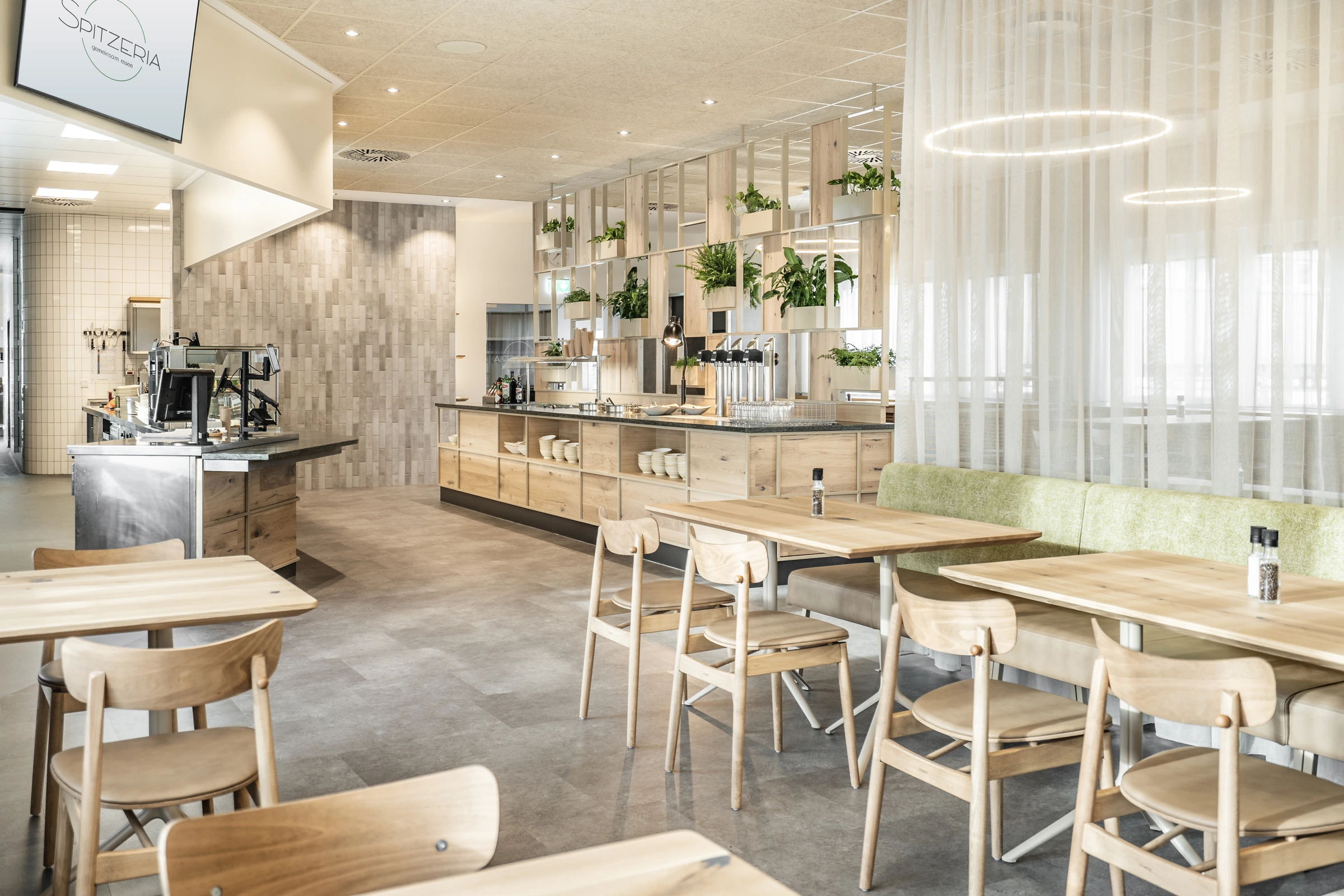 Betriebsrestaurant Spitzeria in Attnang mit Warmhaltestrahler, Tellern auf der Theke und bepflanztem Holzregal im Hintergrund.
