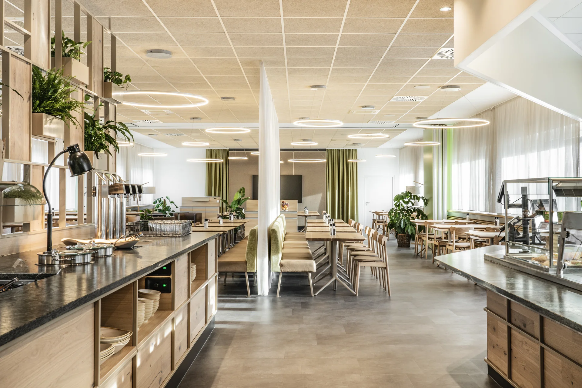 Gesamtansicht des Betriebsrestaurants Spitzeria in Attnang mit Buffettheke, Holzregal, Pflanzen und runden Ringlampen.