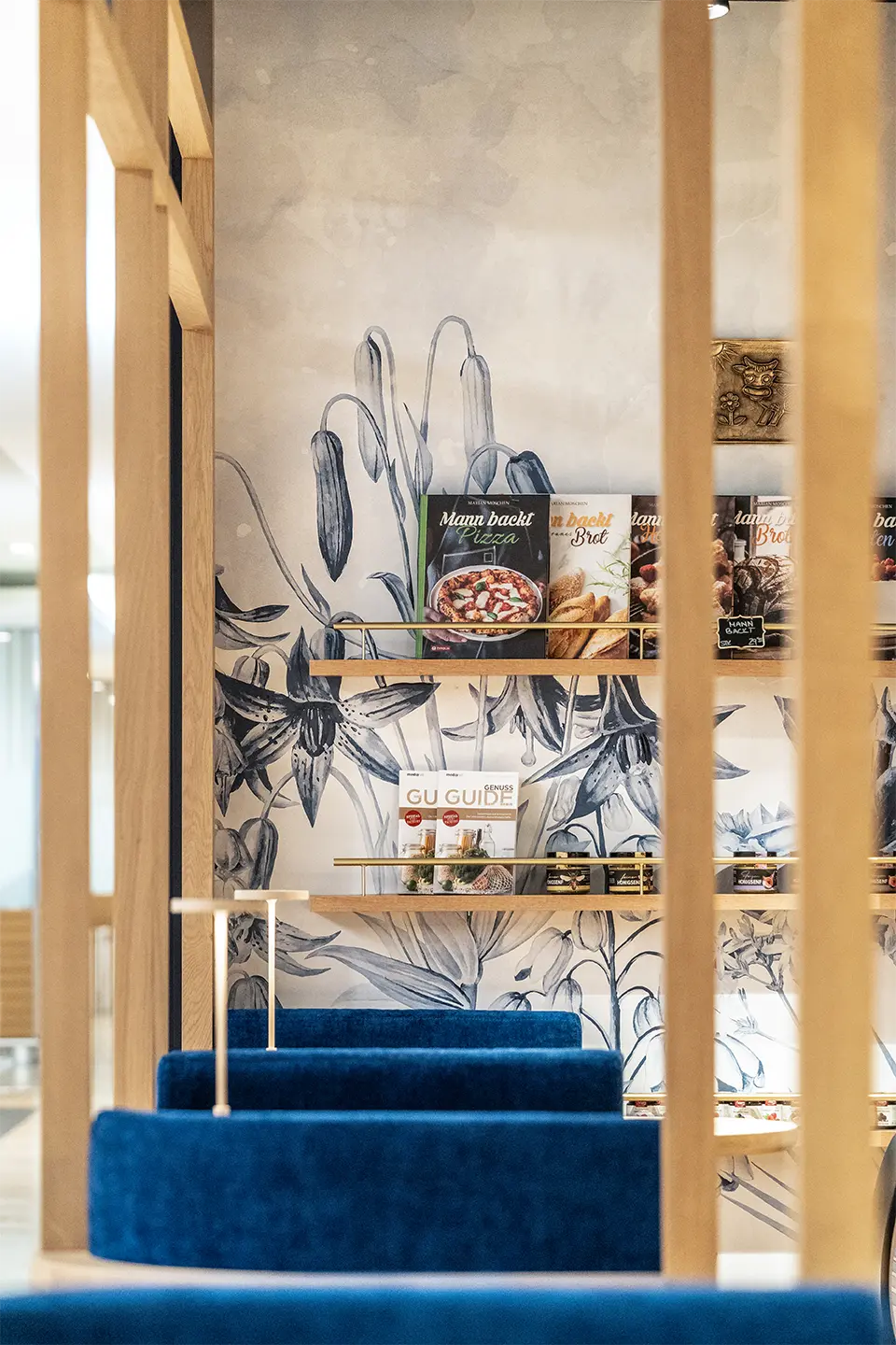 Ein Detailblick in ein Feinkostgeschäft zeigt eine künstlerisch gestaltete Wand mit floralen Motiven, schmale Holzregale mit Kochbüchern und im Vordergrund die Oberkanten von blauen Samtsitzen hinter einer Holzlamellenstruktur.