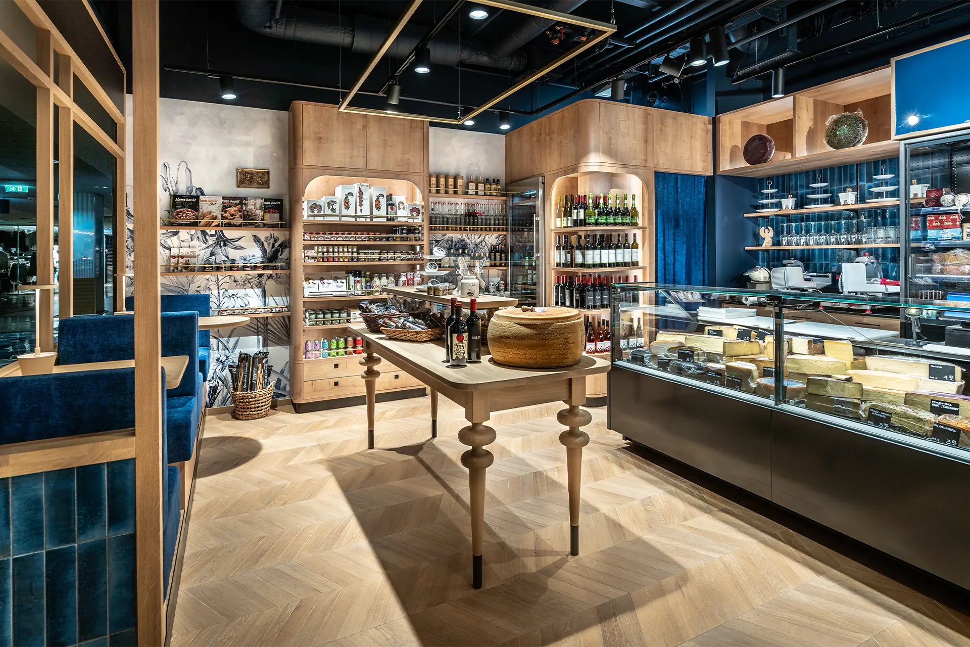 Der Innenraum besticht durch ein hochwertiges Interieur mit Fischgrätparkett, einer eleganten Käsetheke aus Glas und maßgefertigten Holzregalen, die eine exklusive Auswahl an Weinen und Spezialitäten präsentieren.