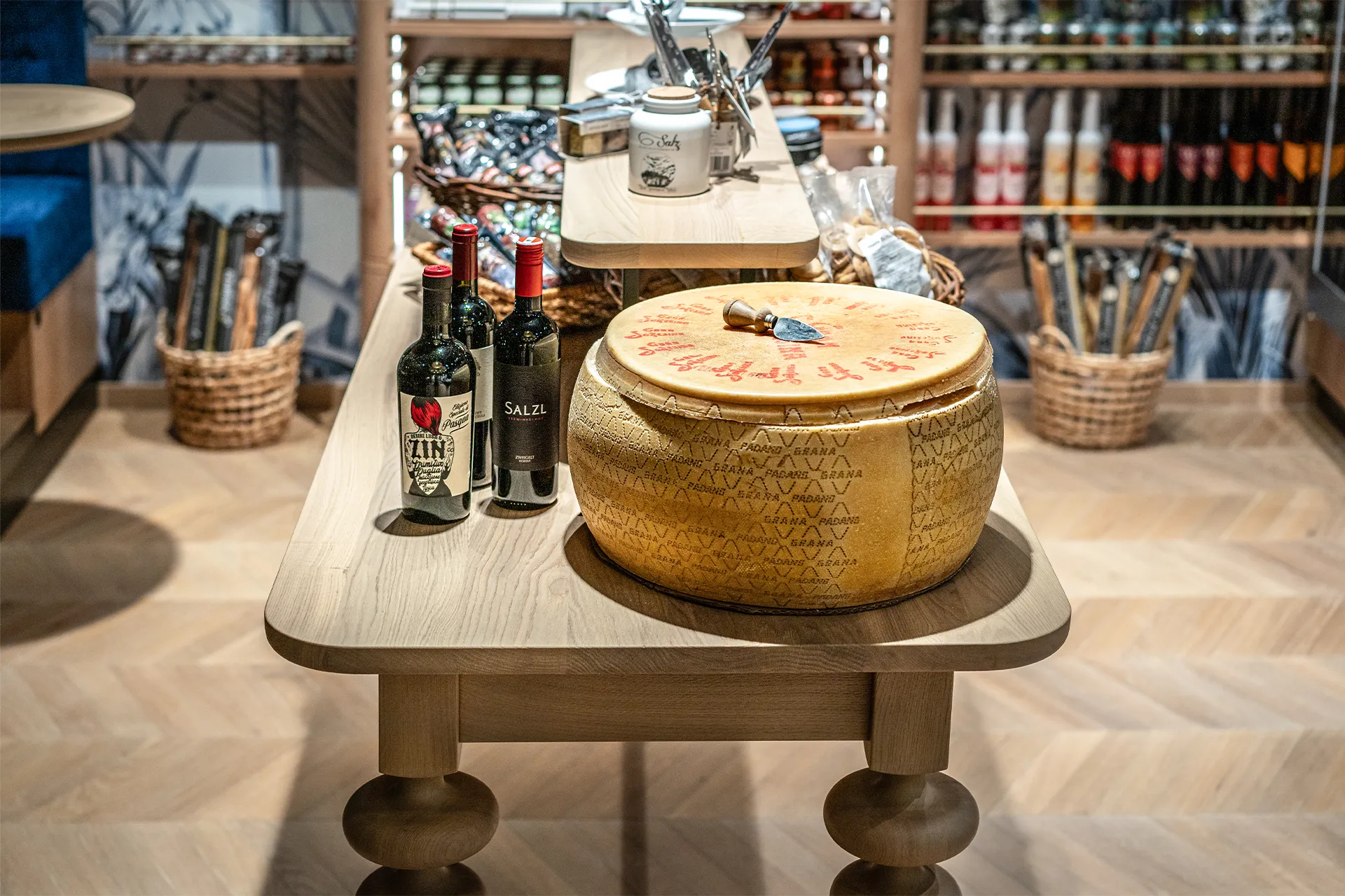 Ein großer Laib Grana Padano und drei Flaschen Rotwein sind ansprechend auf einem rustikalen Holztisch inmitten eines modern gestalteten Feinkostgeschäfts präsentier.