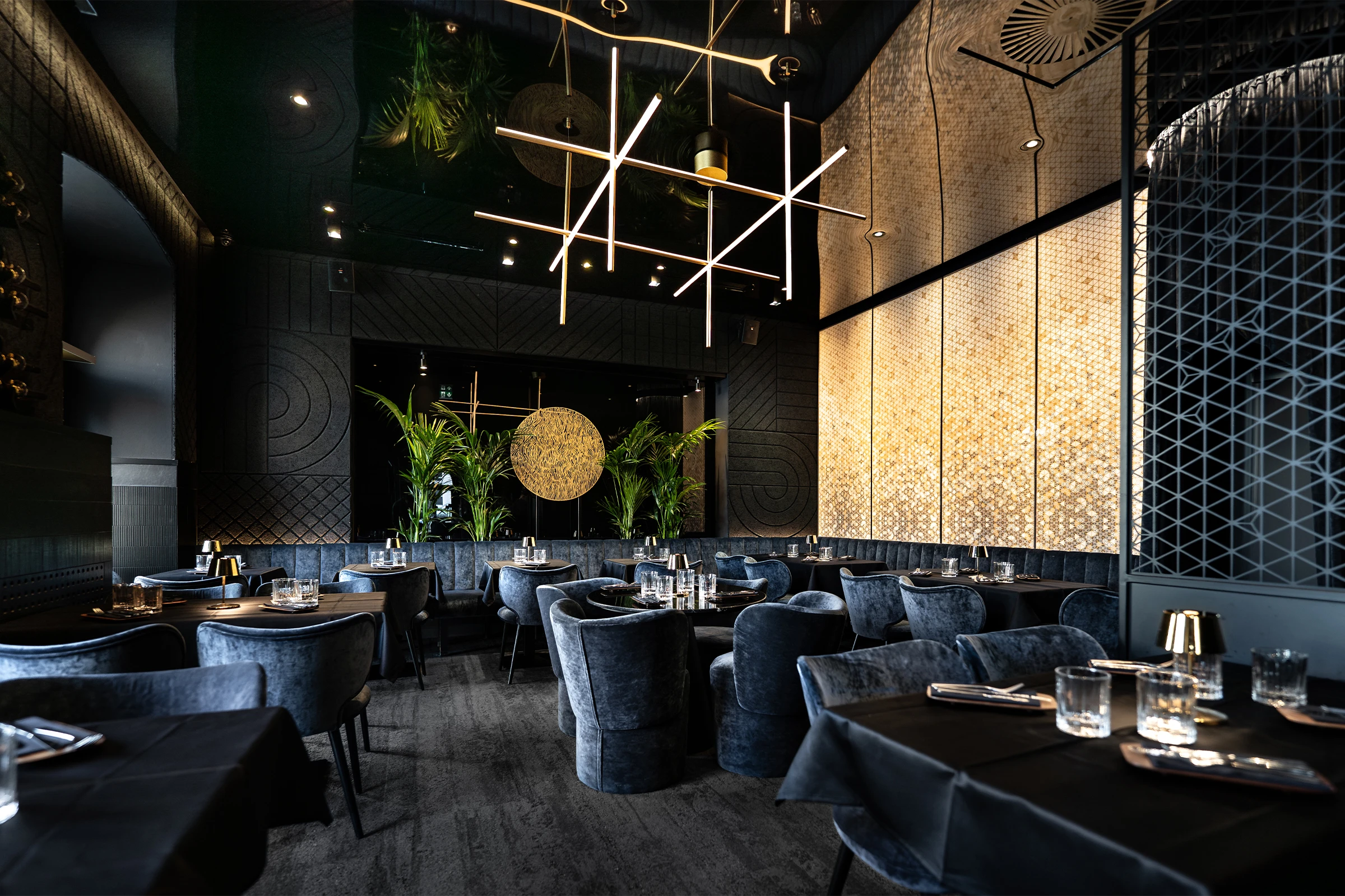 Eleganter Restaurantsaal mit dunkelblauem Samtmobiliar, goldenen Stablampen und einer hinterleuchteten Wandverkleidung in warmem Bernsteinton.