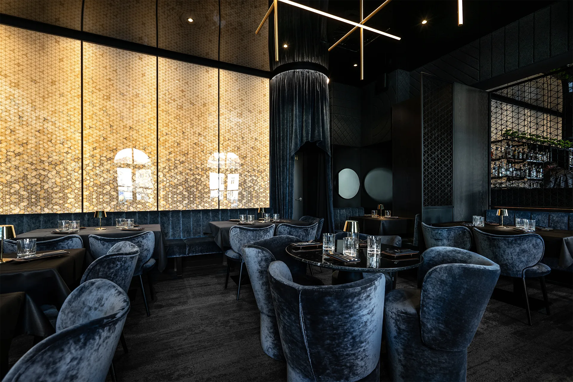 Großzügiger Restaurantraum mit hinterleuchteter Goldwand, blauem Samtmobiliar, einem drapierten Säulenvorhang und geometrischen Metall-Raumteilern.