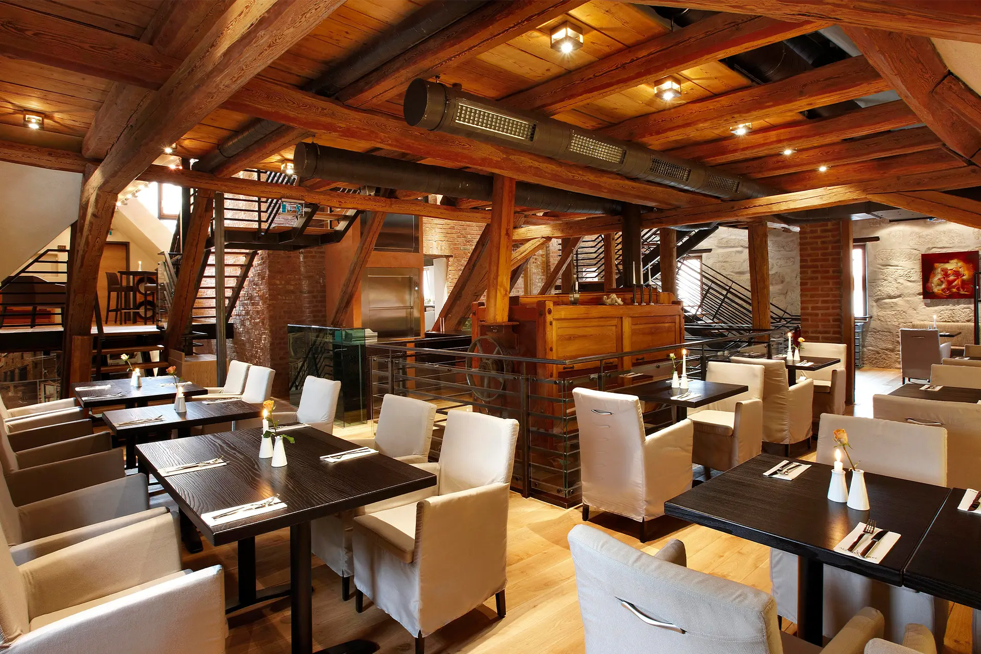 Rustikaler Restaurantsaal mit weiß gepolsterten Stühlen an dunklen Holztischen unter einem imposanten Holzbalkengebälk mit sichtbaren Industrierohren und Backsteinwänden.