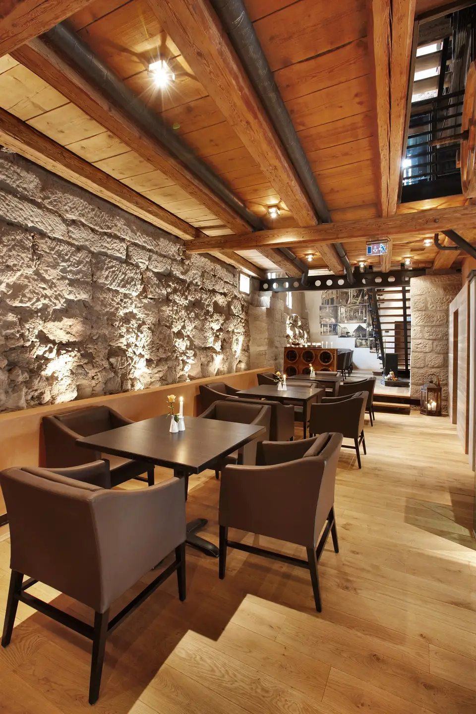 Restaurantbereich der Alten Mälzerei mit beleuchteter Natursteinwand, Holzbalkendecke, gedeckten Tischen und braunen Sesseln auf hellem Eichenparkett.