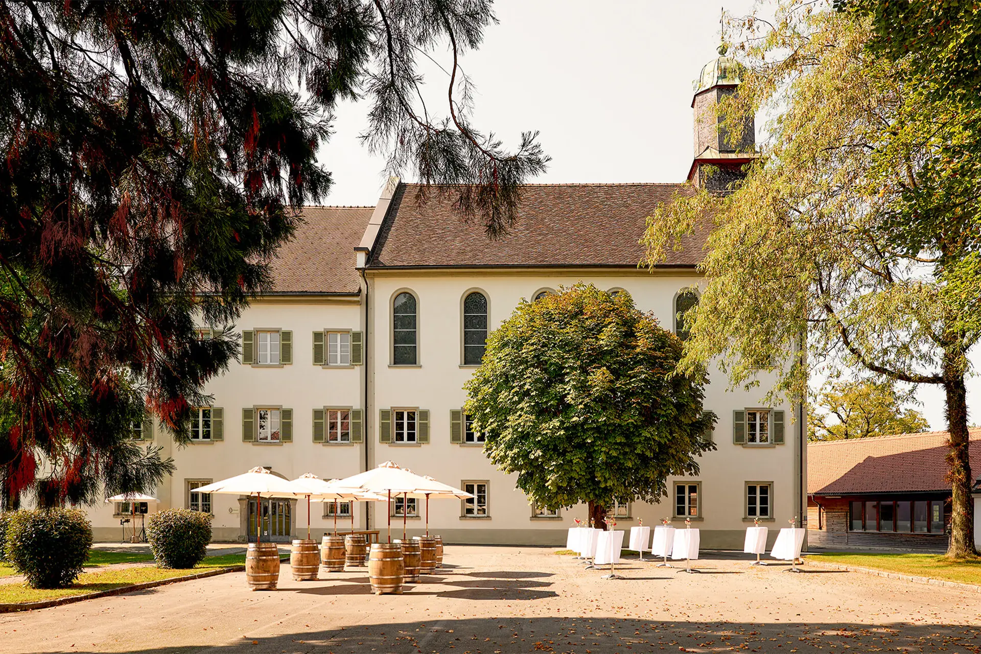 Historisches Gebäude der Aula Bernardi mit gepflegtem Vorplatz, auf dem Weinfässer als Stehtische und weiße Cocktailtische für einen Empfang im Freien bereitstehen.