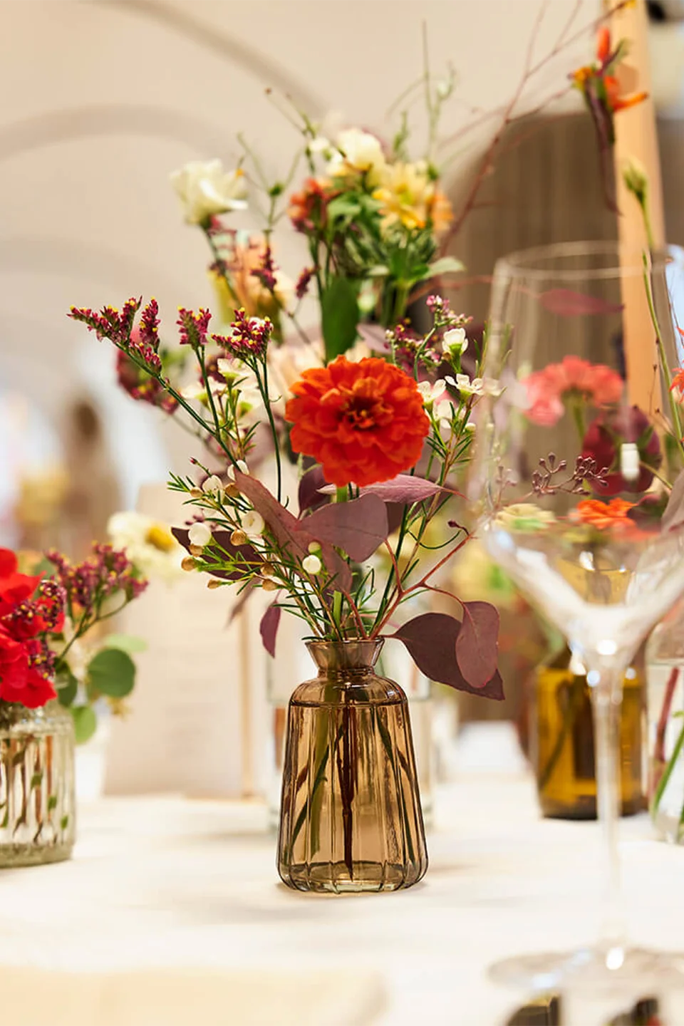 Kleine braune Glasvase mit einem lockeren Gesteck aus oranger Dahlie, Wiesenblumen und burgunderroten Blättern als Tischdekoration im Gewölbesaal.