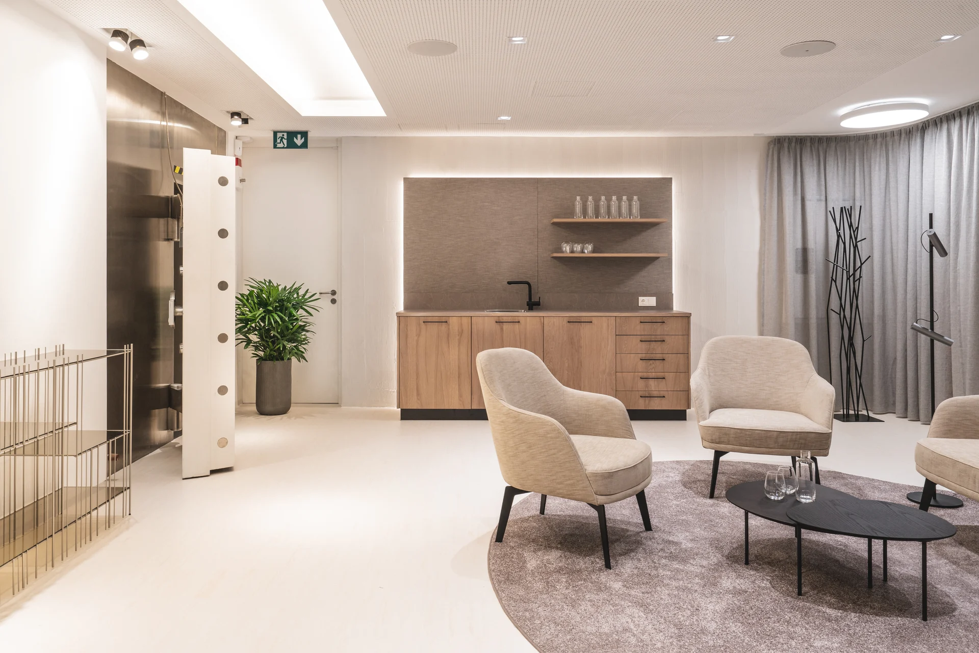 Großzügiger Loungebereich bei Biogena Wien mit beigen Sesseln, Rundteppich, beleuchteter Holzküche und Blick auf die hyperbare Kammer im Hintergrund