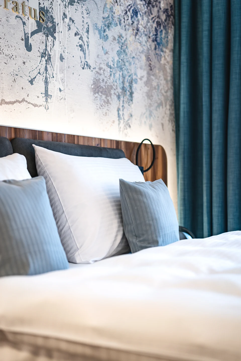 Detailansicht des Bettes im Hotel Blauer Bock Passau mit blauen Dekokissen, weißer Bettwäsche, Holzkopfteil und kunstvoller Wandtapete mit türkisem Vorhang.