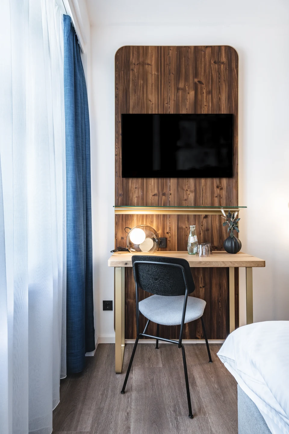 Schreibtischbereich im Hotel Blauer Bock Passau mit dunkler Holzvertäfelung, wandmontiertem Fernseher, goldenem Schreibtisch, modernem Stuhl und blauem Vorhang.