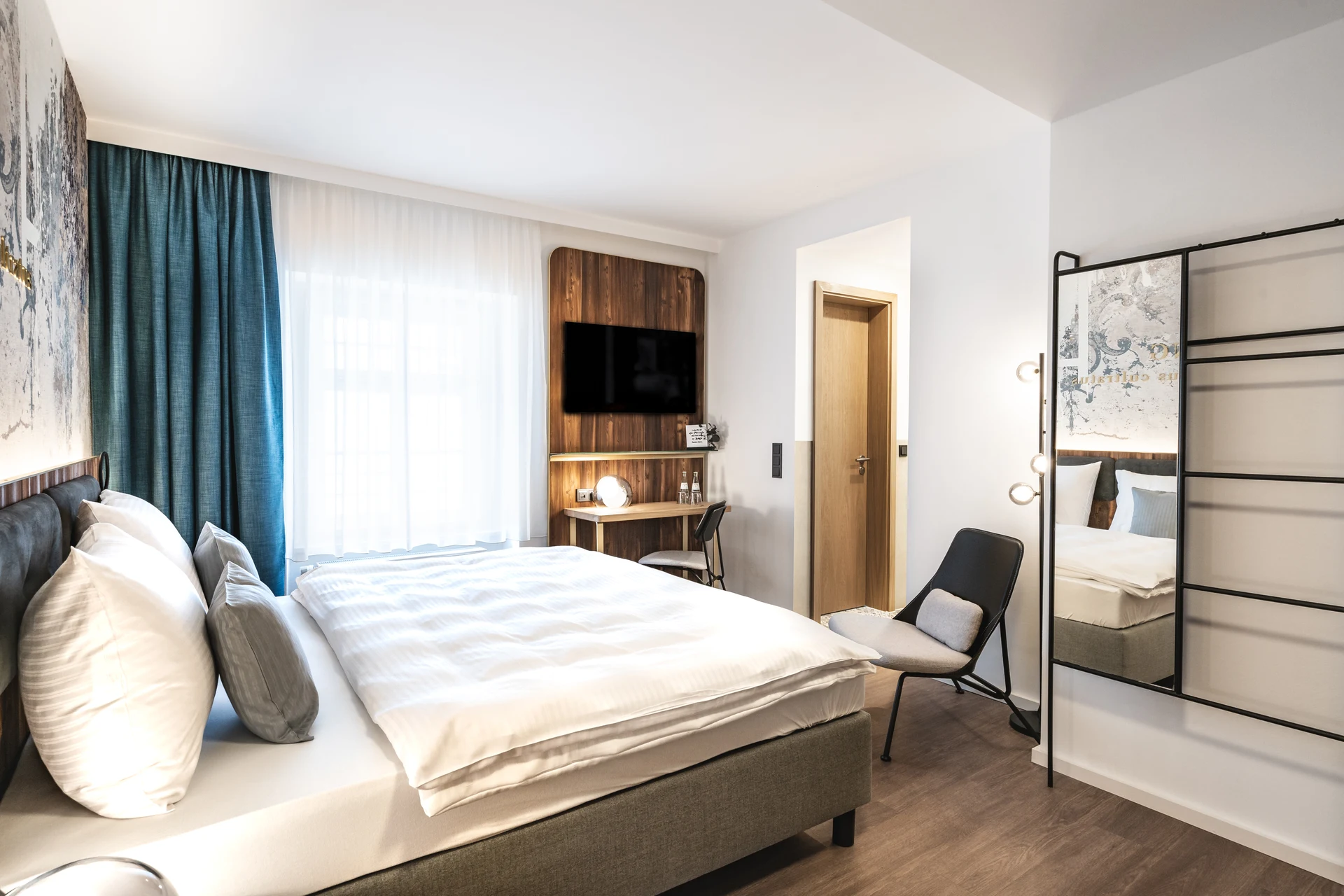 Helles Hotelzimmer im Blauer Bock Passau mit Boxspringbett, türkisem Vorhang, Holz-TV-Paneel, Schreibtisch und industriellem Spiegelrahmen.