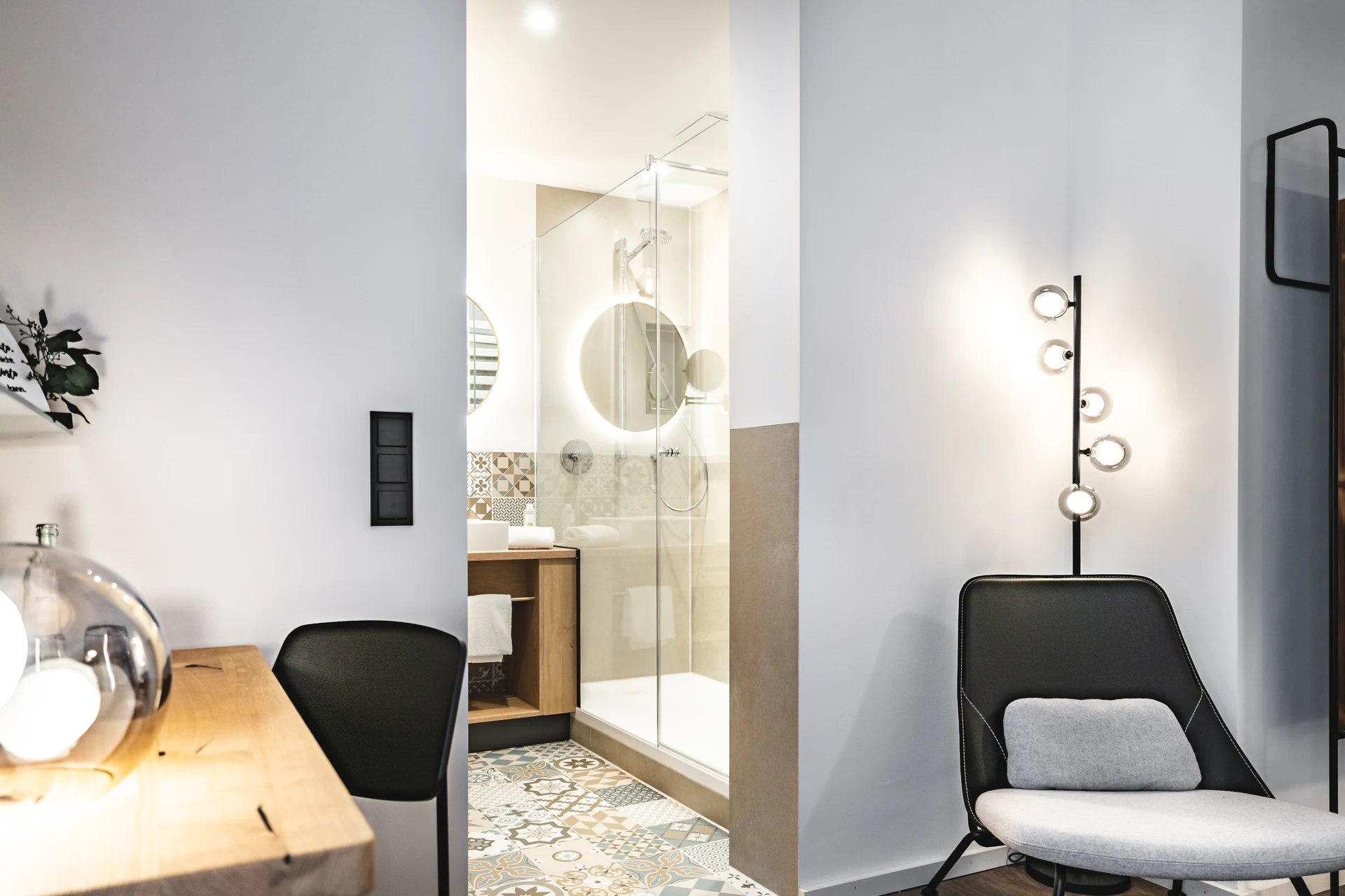 Hotelzimmer im Blauer Bock Passau mit Blick ins Bad mit Glasdusche und Mosaikfliesenboden, schwarzem Loungesessel mit Kugelleuchte und Schreibtischbereich.