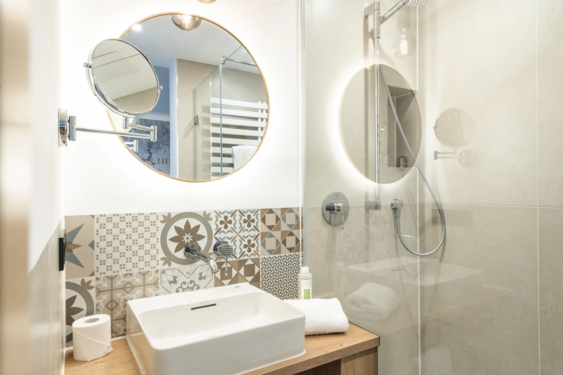 Modernes Badezimmer im Hotel Blauer Bock Passau mit ornamentalen Patchwork-Kacheln, rundem Goldrahmen-Spiegel, wei&szlig;em Aufsatzwaschbecken auf Holzwaschtisch und ebenerdiger Glasdusche.