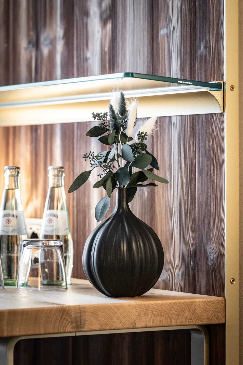 Detailaufnahme im Hotel Blauer Bock Passau mit schwarzer Keramikvase, Trockenblumen und Eukalyptus auf einem Holzregal mit Messinghalterung, daneben Glasflaschen und Trinkgläser vor einer Altholzwand.