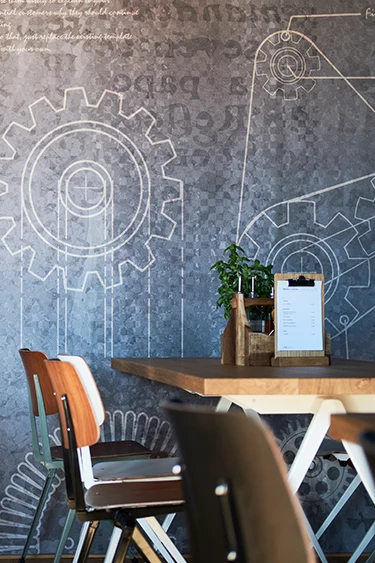 Industriell gestaltete Wandgestaltung mit aufgezeichneten Zahnrädern und Schriftmotiven hinter einem schlicht gedeckten Restauranttisch mit Holzstühlen