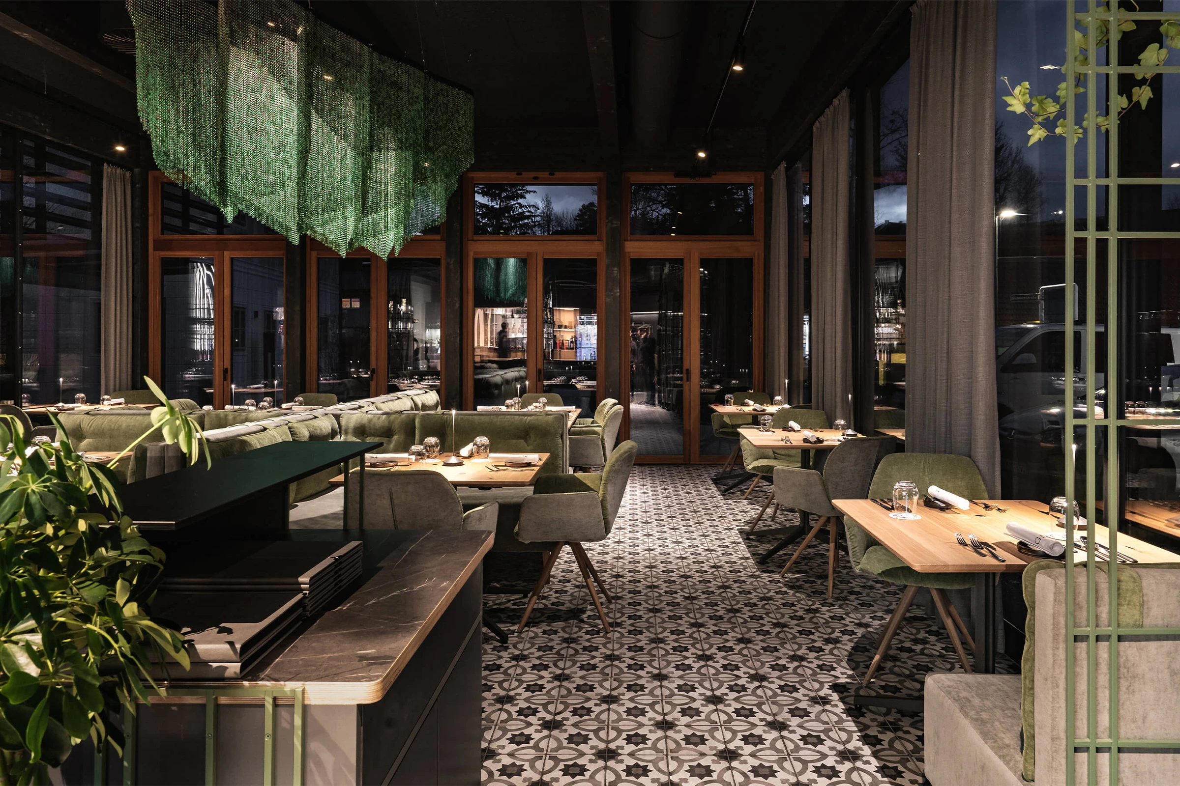 Ein elegant eingerichteter Restaurant-Gastraum mit gemusterten Bodenfliesen, grünen Polstermöbeln und einer großen, wellenförmigen Skulptur aus grünen Ketten an der Decke.