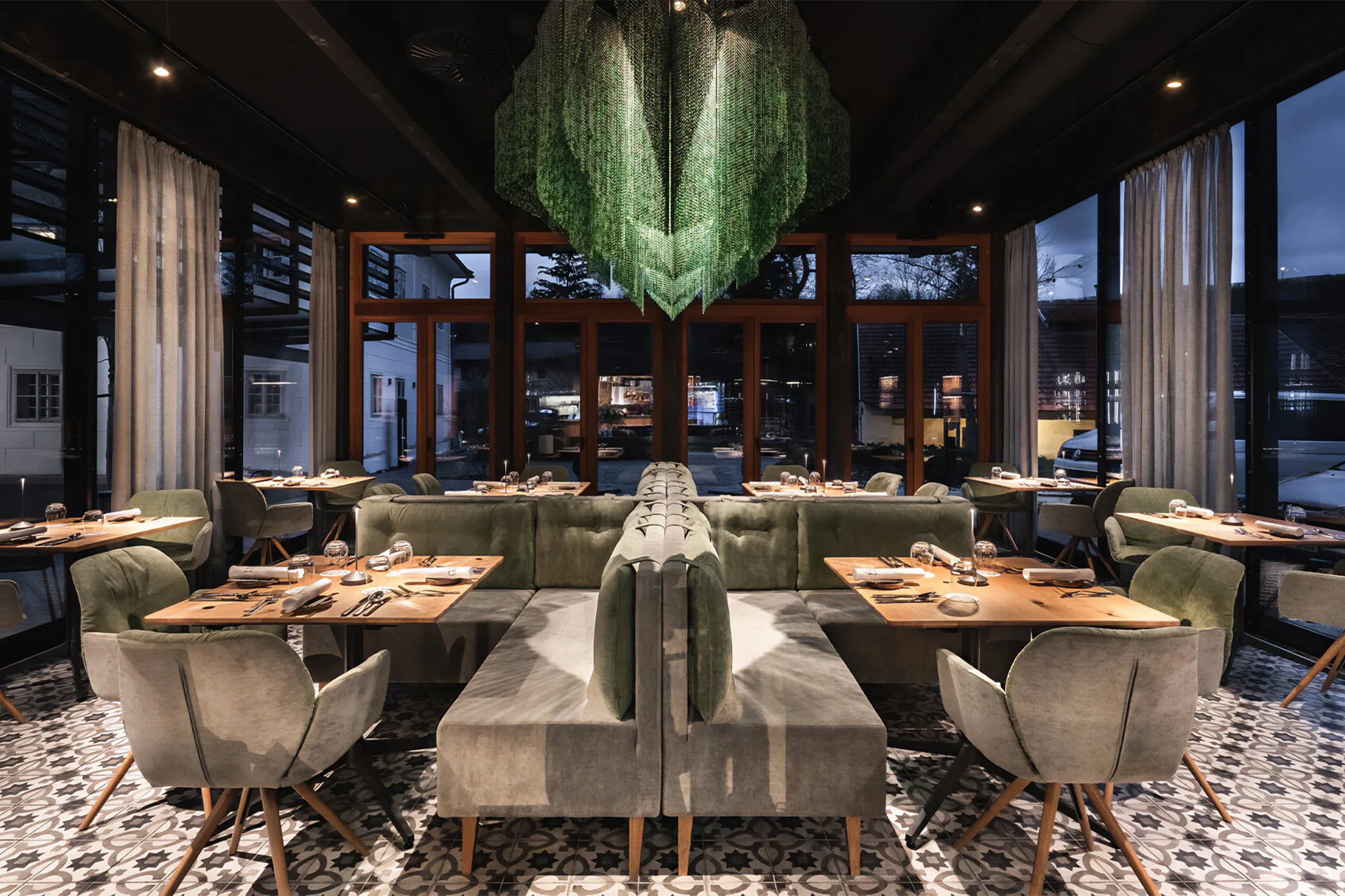 Der modern gestaltete Gastraum eines Fine-Dining-Restaurants mit kreuzförmig angeordneten, olivgrünen und grauen Sitzbänken und Tischen vor großen Panoramafenstern.
