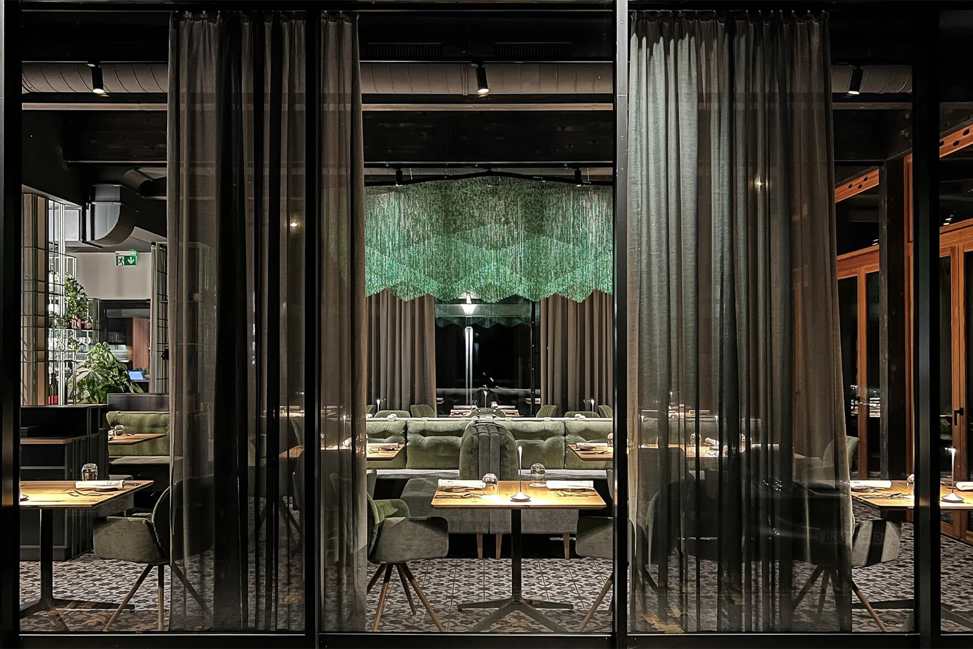 Blick von außen durch eine große Fensterfront mit dunklen Vorhängen auf den elegant beleuchteten Sitzbereich eines modernen Restaurants.