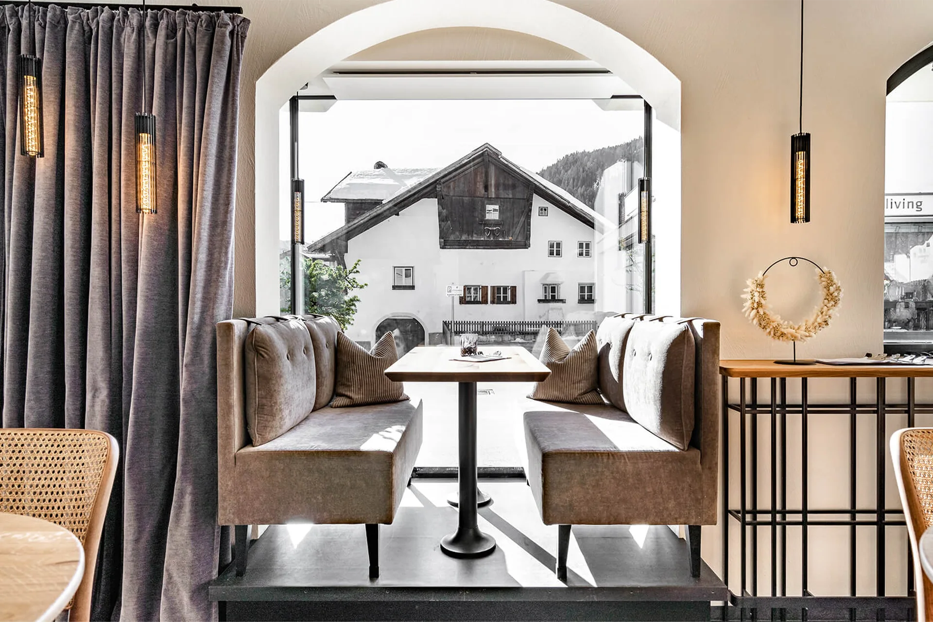 Ein gemütlicher Fensterplatz mit einer grau-beigen Polsterbank, hellem Holztisch und einem Vorhang.