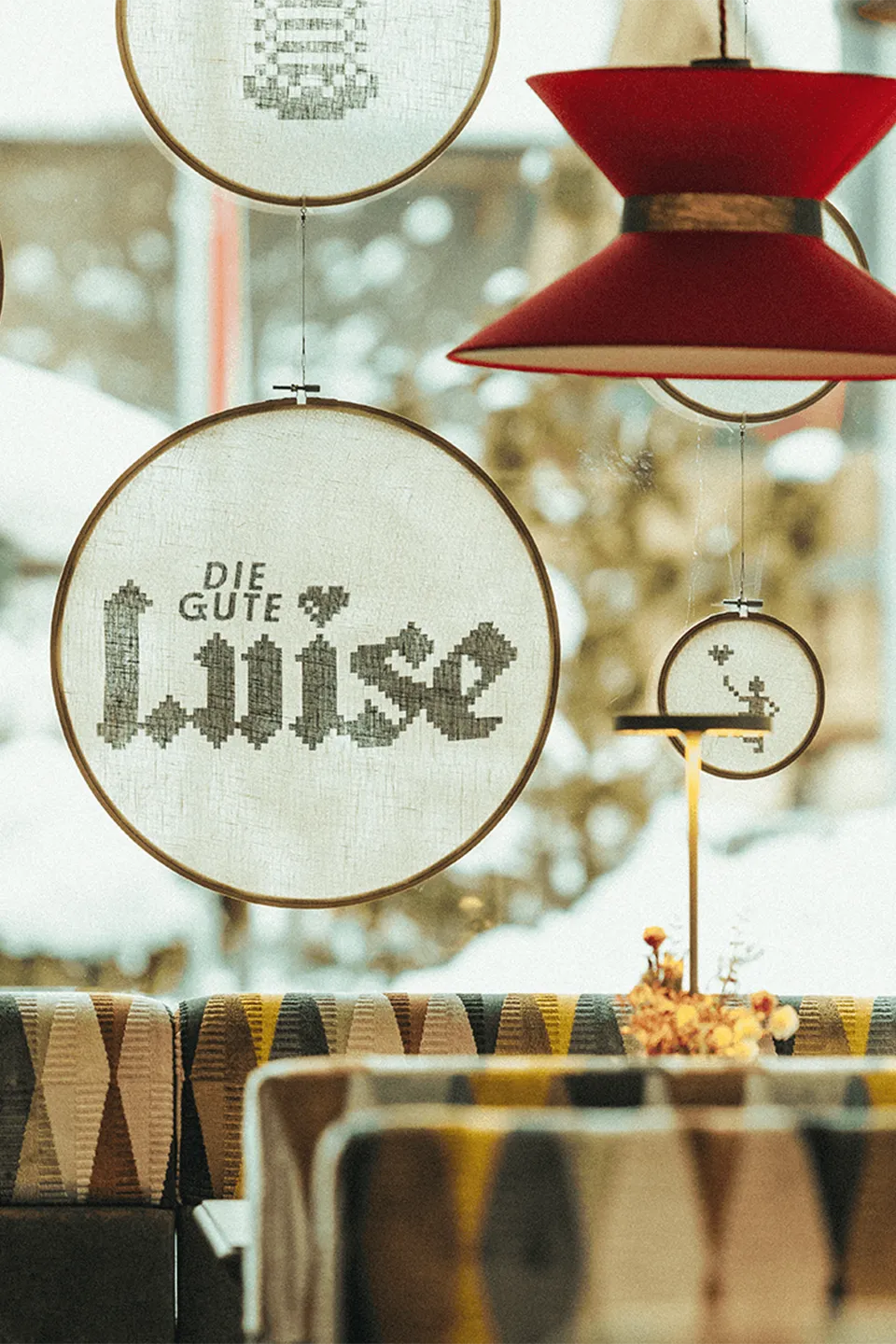 Vor einem Fenster hängende Stickrahmen, in die der Schriftzug und Logo „Die gute Luise“ und Pixelgrafiken eingespannt ist.