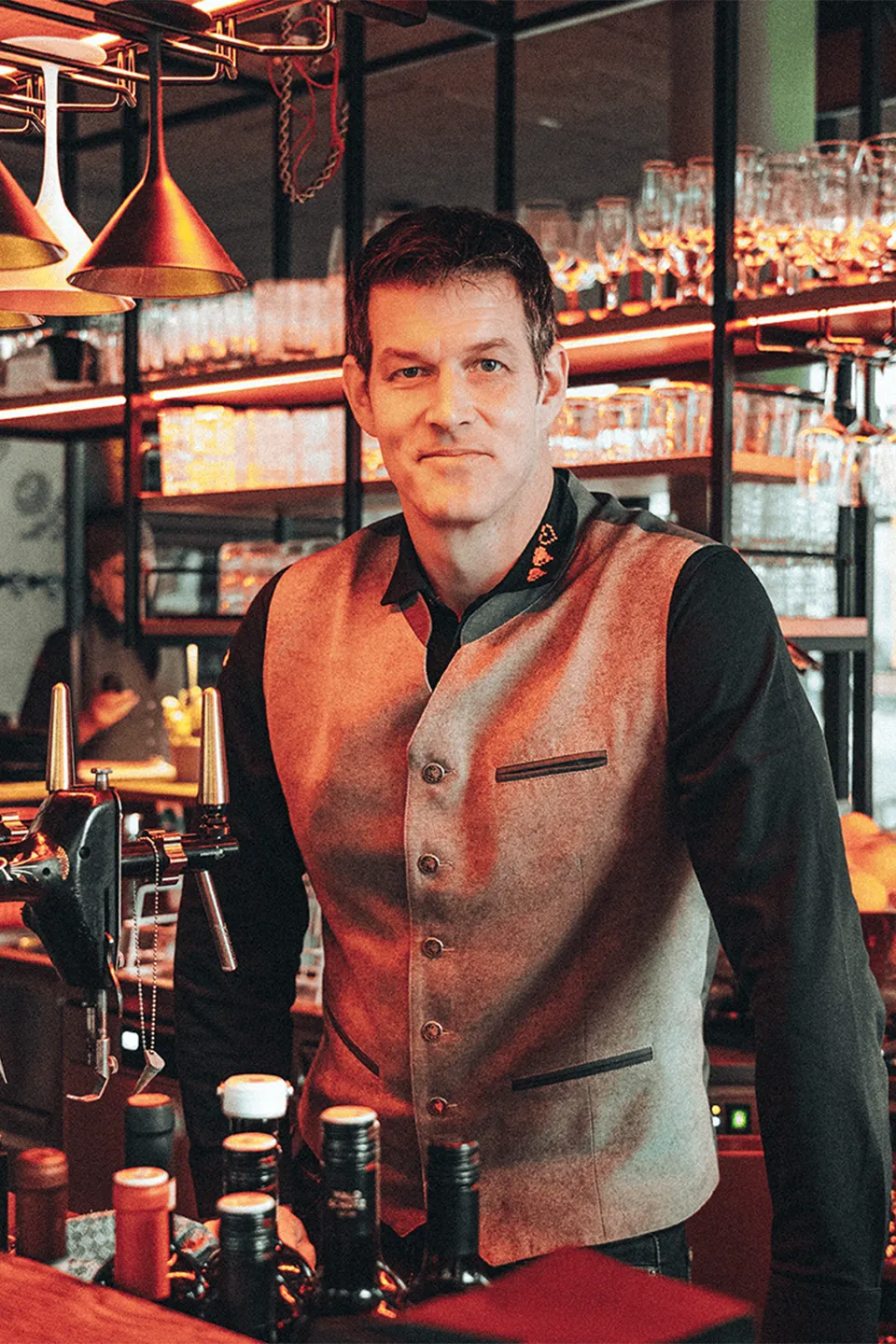 Ein sympathisch lächelnder Geschäftsführer, der in mitten der freistehenden Bar im Lokal steht und in die Kamera sieht.