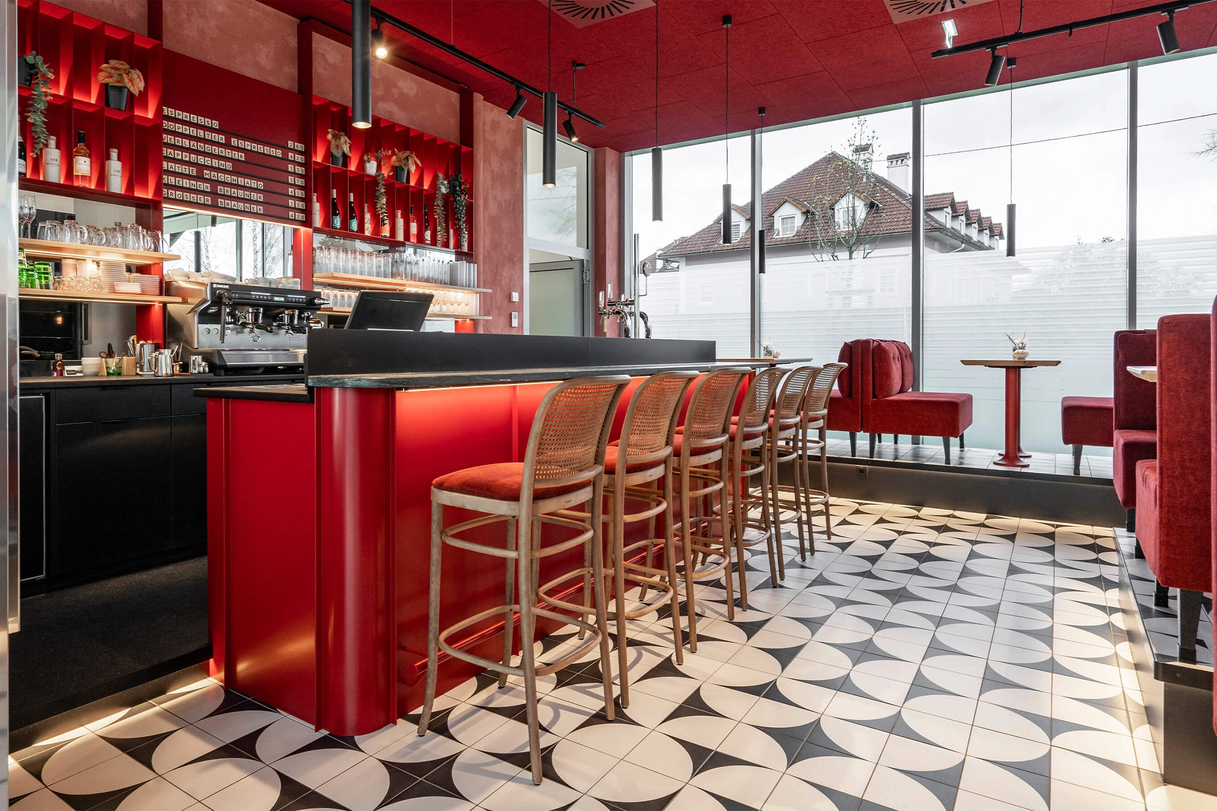 Der Innenraum eines modernen Cafés mit einer roten Theke, Barhockern aus Holz und Geflecht, einem schwarz-weiß gemusterten Fliesenboden und großen Fensterfronten.
