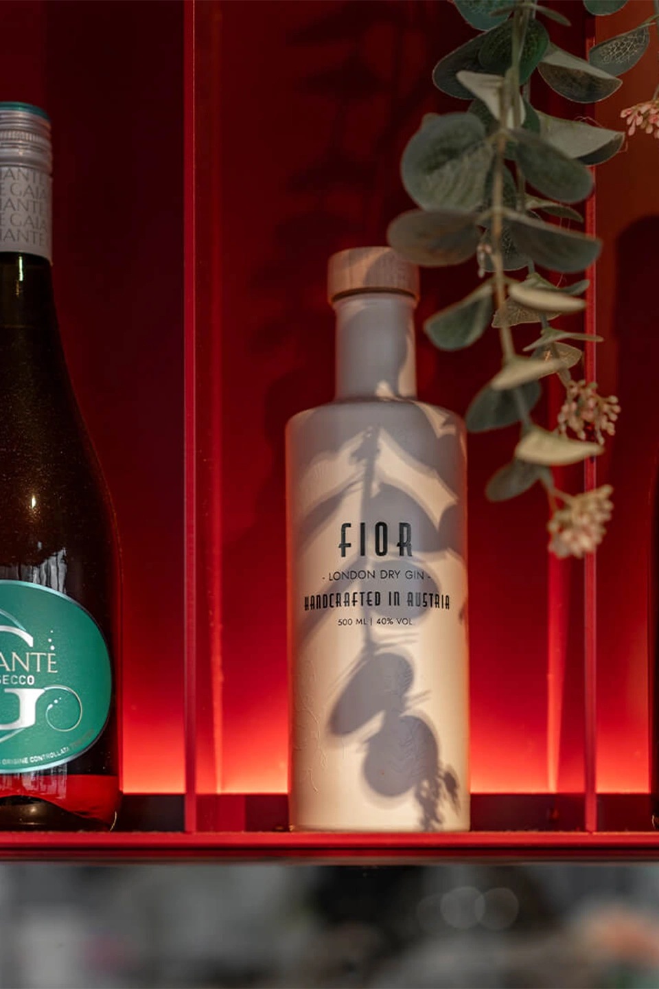 Eine Nahaufnahme eines roten Metallregals mit Flaschen, auf dessen Rückwand der Schatten einer Pflanze fällt, darunter eine weiße Flasche mit der Aufschrift „FIOR London Dry Gin“.