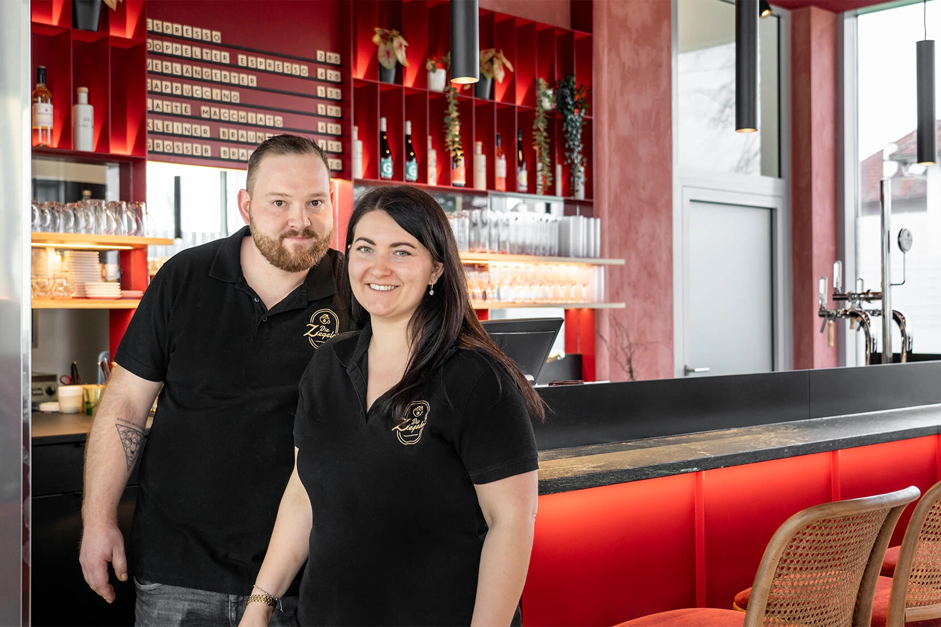 Ein Mann und eine Frau, die Gesch&auml;ftsf&uuml;hrer, in schwarzen Poloshirts stehen l&auml;chelnd hinter der roten Theke des Caf&eacute;s.