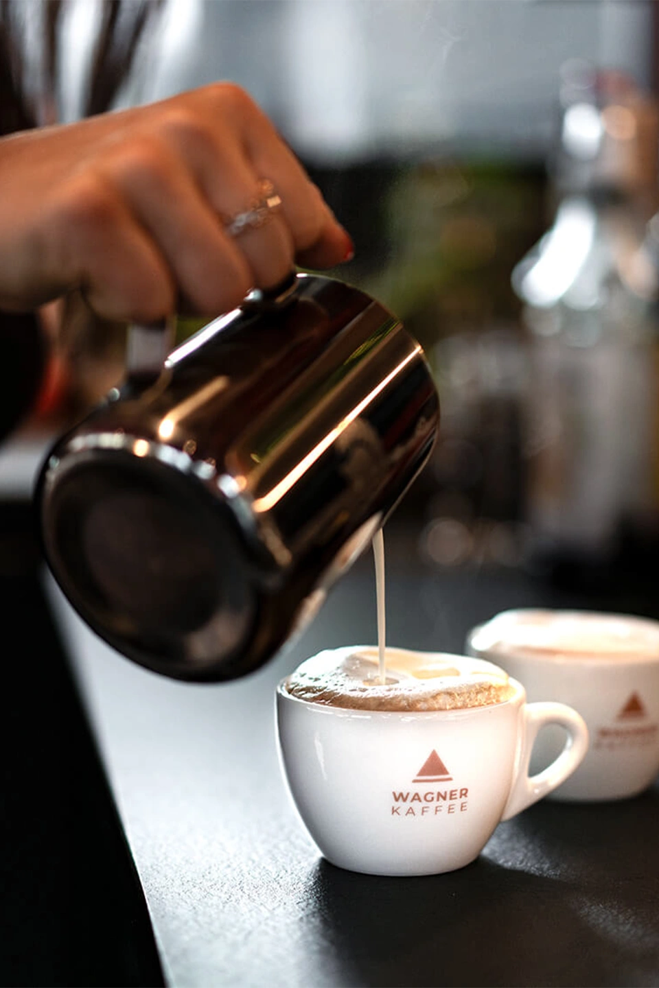 Eine Hand gießt aufgeschäumte Milch aus einem Edelstahlkännchen in eine weiße Kaffeetasse mit dem Logo „WAGNER KAFFEE“.