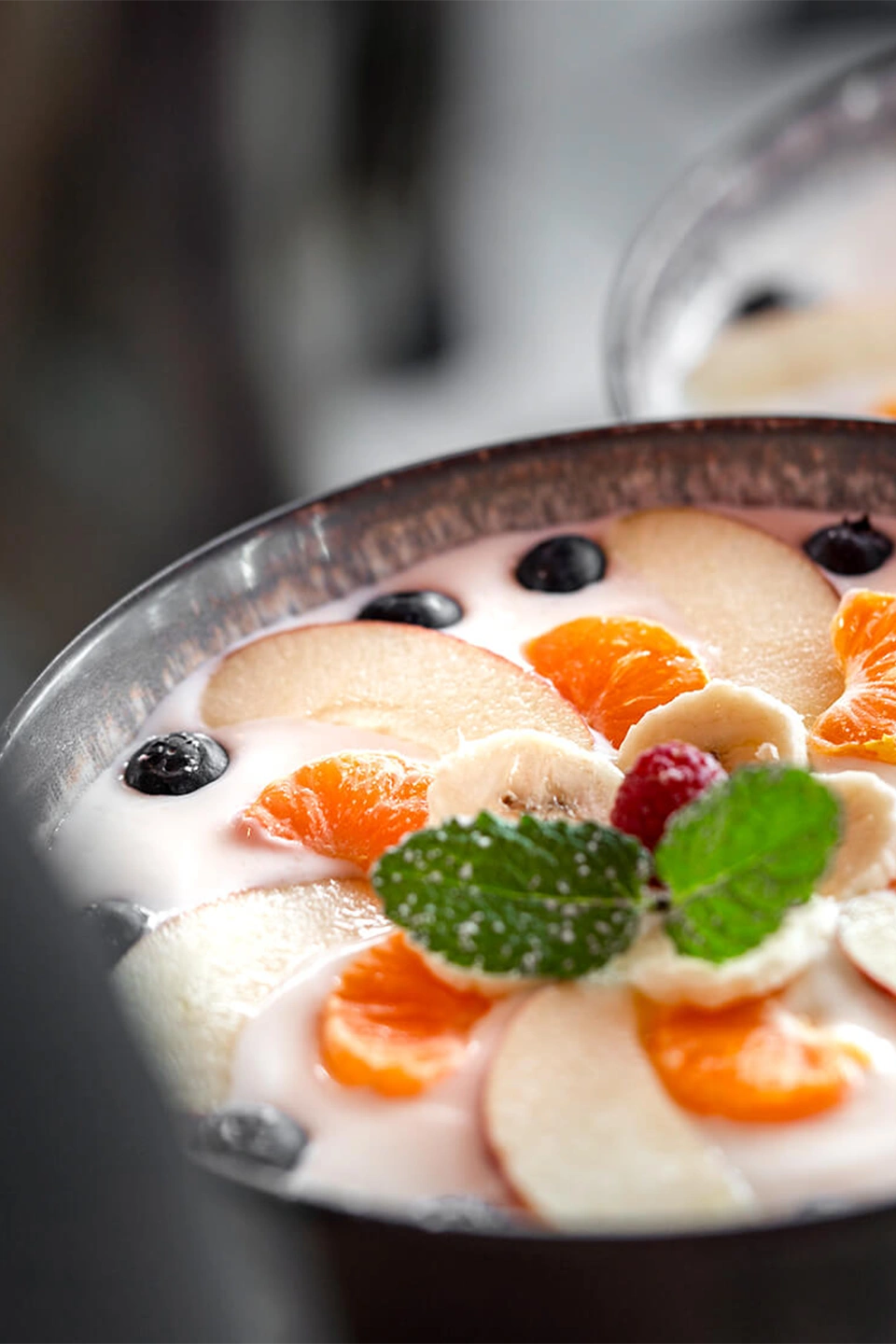 Eine Nahaufnahme einer Frühstücksschale mit Joghurt, garniert mit Apfelscheiben, Mandarinensegmenten, Bananenscheiben, Blaubeeren, einer Himbeere und frischer Minze.