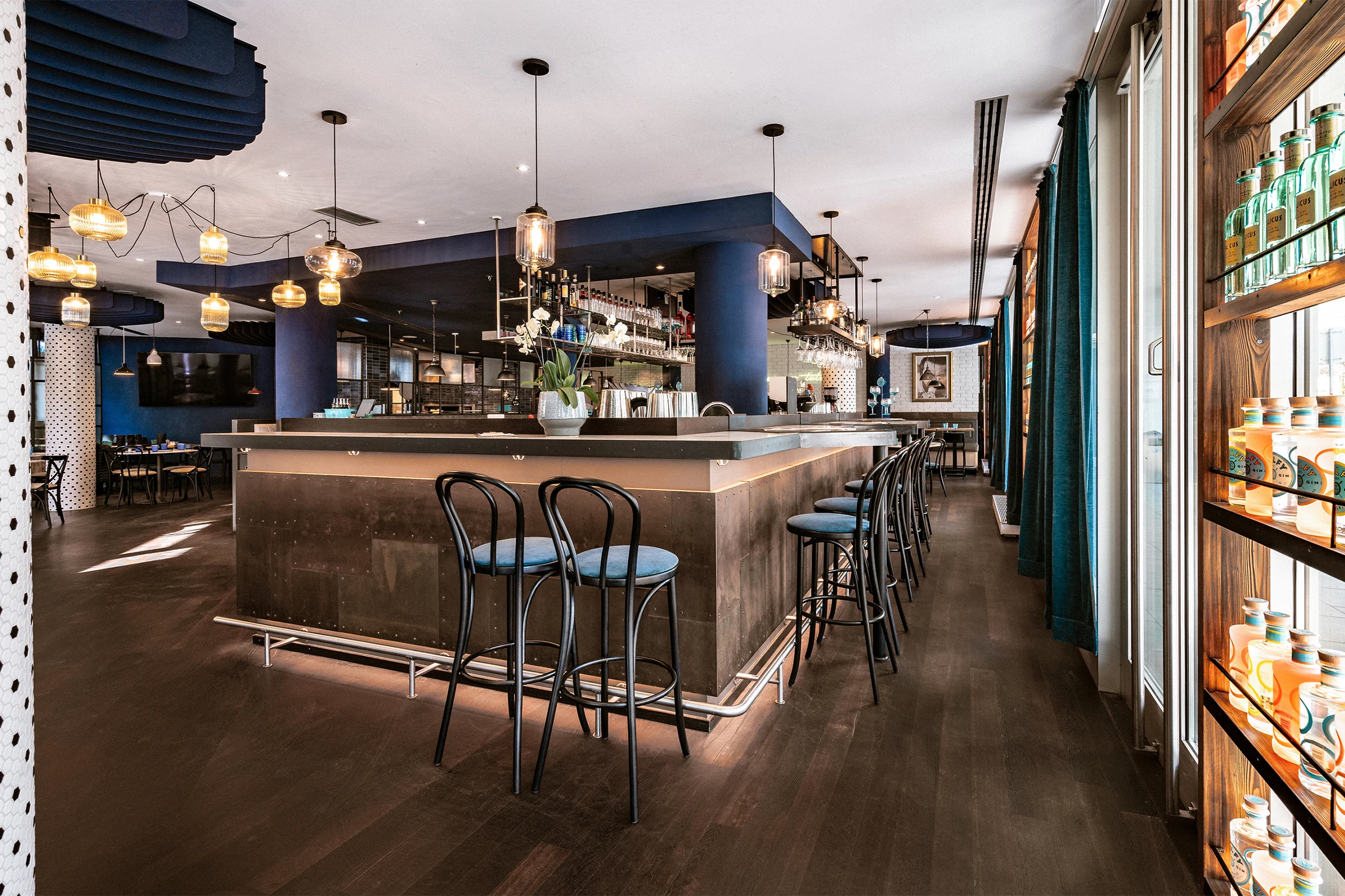 Der weitläufige Innenraum eines Restaurants zeigt eine zentrale Bar mit metallischer Verkleidung, umgeben von dunklen Holzböden und blau abgesetzten Deckenelementen.