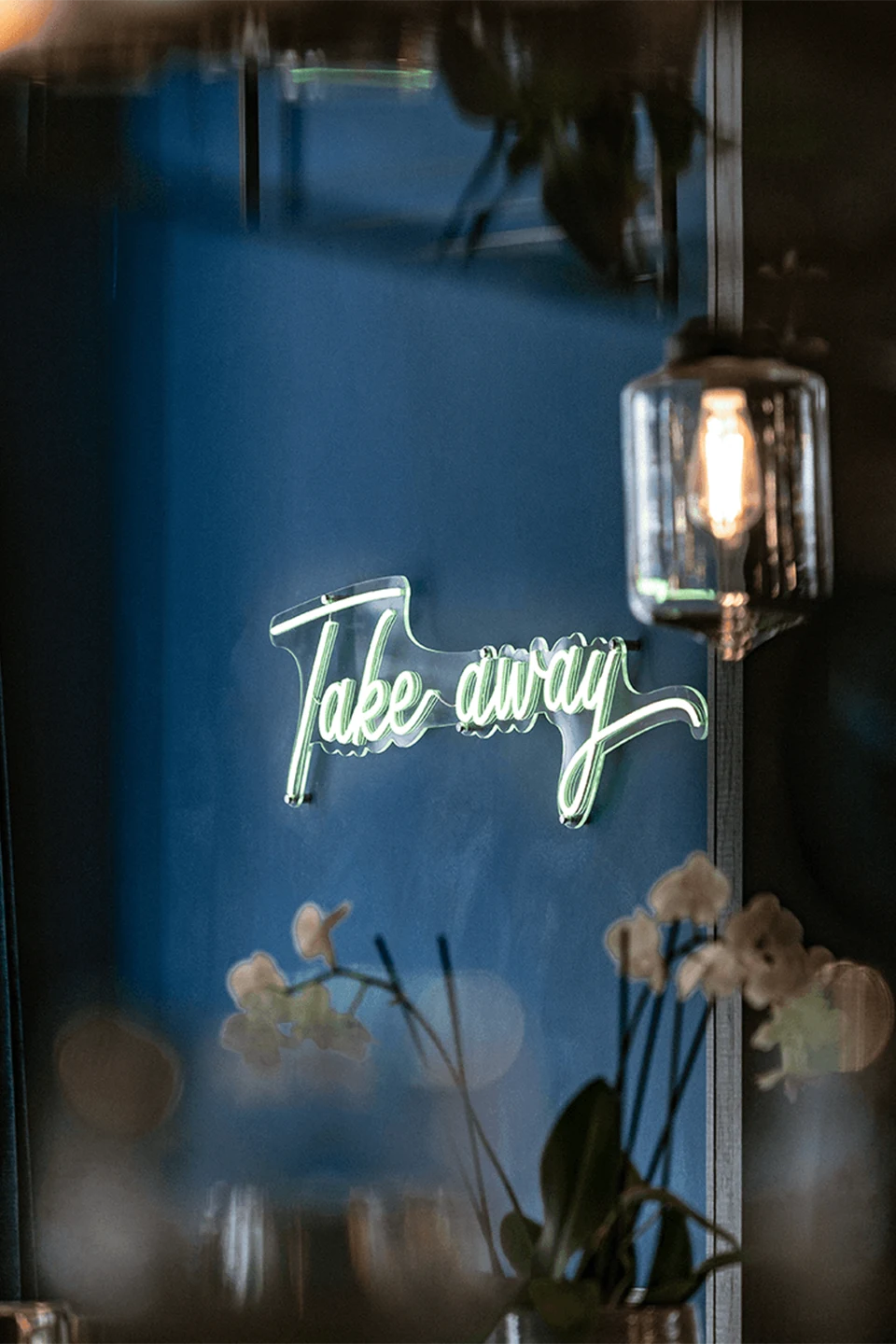 Ein leuchtender, Neonschriftzug mit den Worten „Take away“ hängt an einer dunkelblauen Wand hinter unscharfen Pflanzen im Vordergrund.