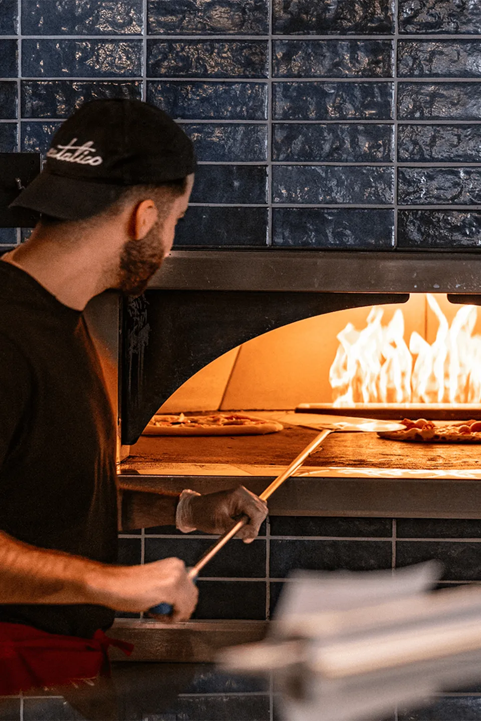 Ein Pizzabäcker mit schwarzer Kappe schiebt mit einer langen Pizzaschaufel eine Pizza in einen lodernden Steinofen.