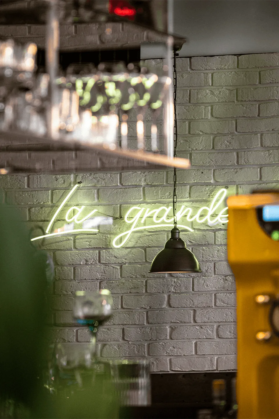 An einer weiß gestrichenen Ziegelwand leuchtet hinter tief hängenden Lampen ein geschwungener Neonschriftzug mit den teilweise sichtbaren Worten „La grande“.