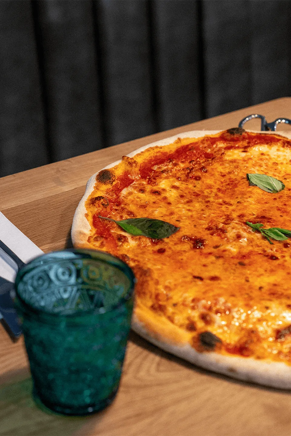 Eine große Pizza mit geschmolzenem Käse und frischen Basilikumblättern liegt auf einem Holztisch neben einem türkisen Strukturglas.