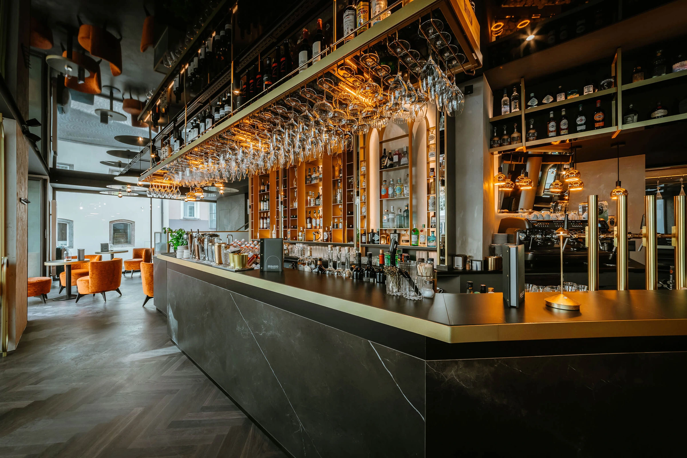 Elegante Bar des Erbario in Schwaz, in Tirol, mit schwarzem Marmortresen, goldenen Akzenten, hängenden Weingläsern und warmem Licht vor dem vollbestückten Flaschenregal.