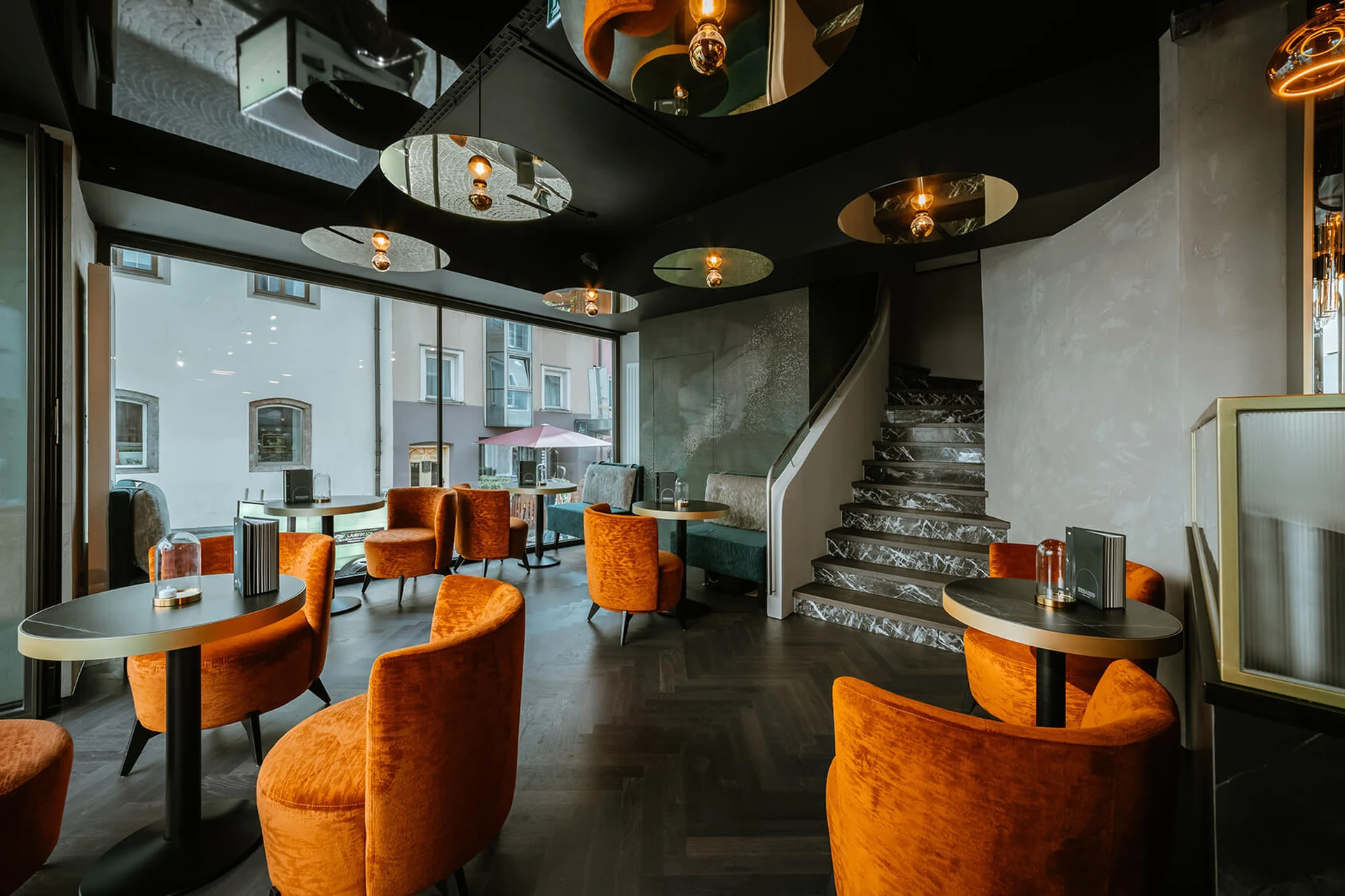 Stimmungsvoller Loungebereich des Erbario in Schwaz mit orangefarbenen Samtsesseln, schwarzer Spiegeldecke, runder Marmortreppe und Blick auf die Altstadt.