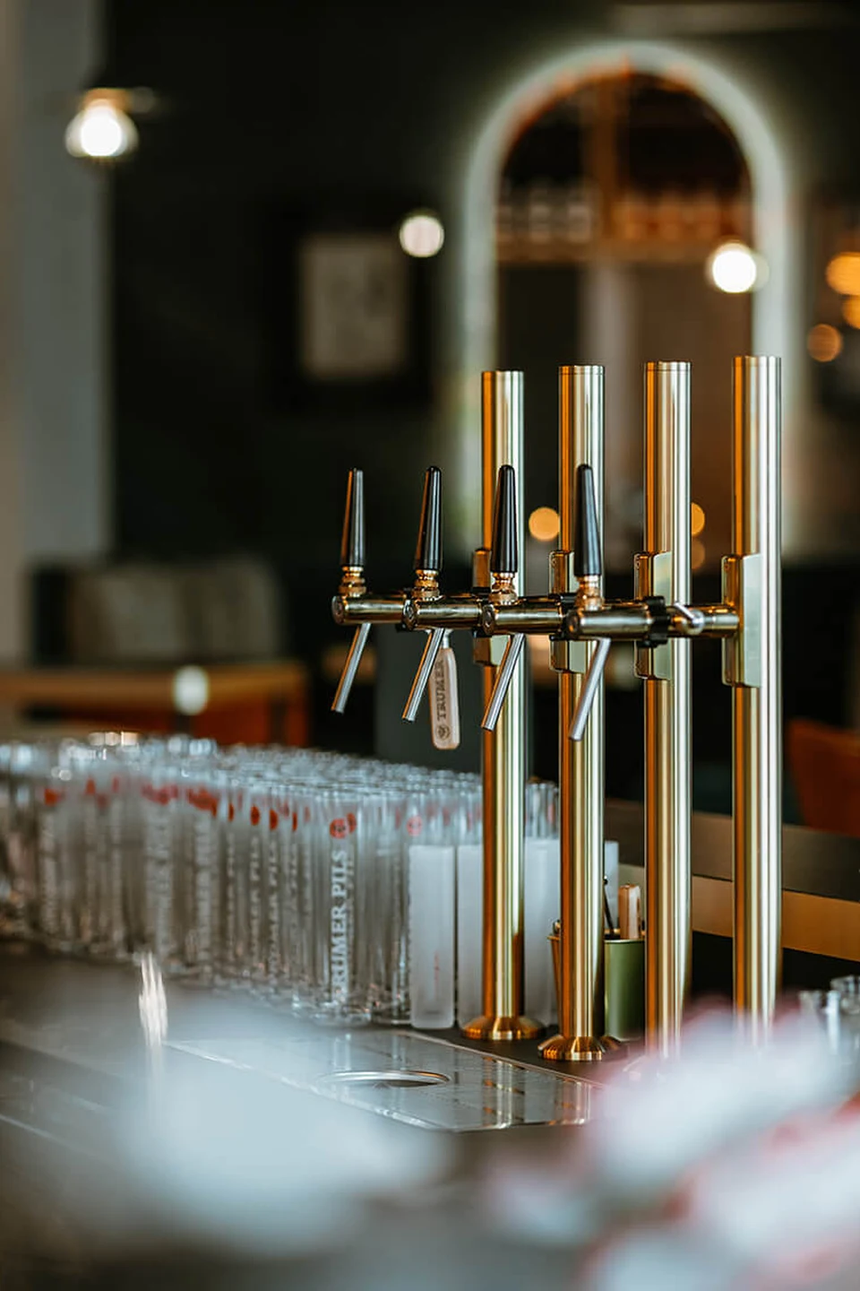 Detailansicht der goldenen Zapfanlage im Erbario in Schwaz mit aufgereihten Biergläsern auf dem Marmortresen und Bogendurchgang im Hintergrund.