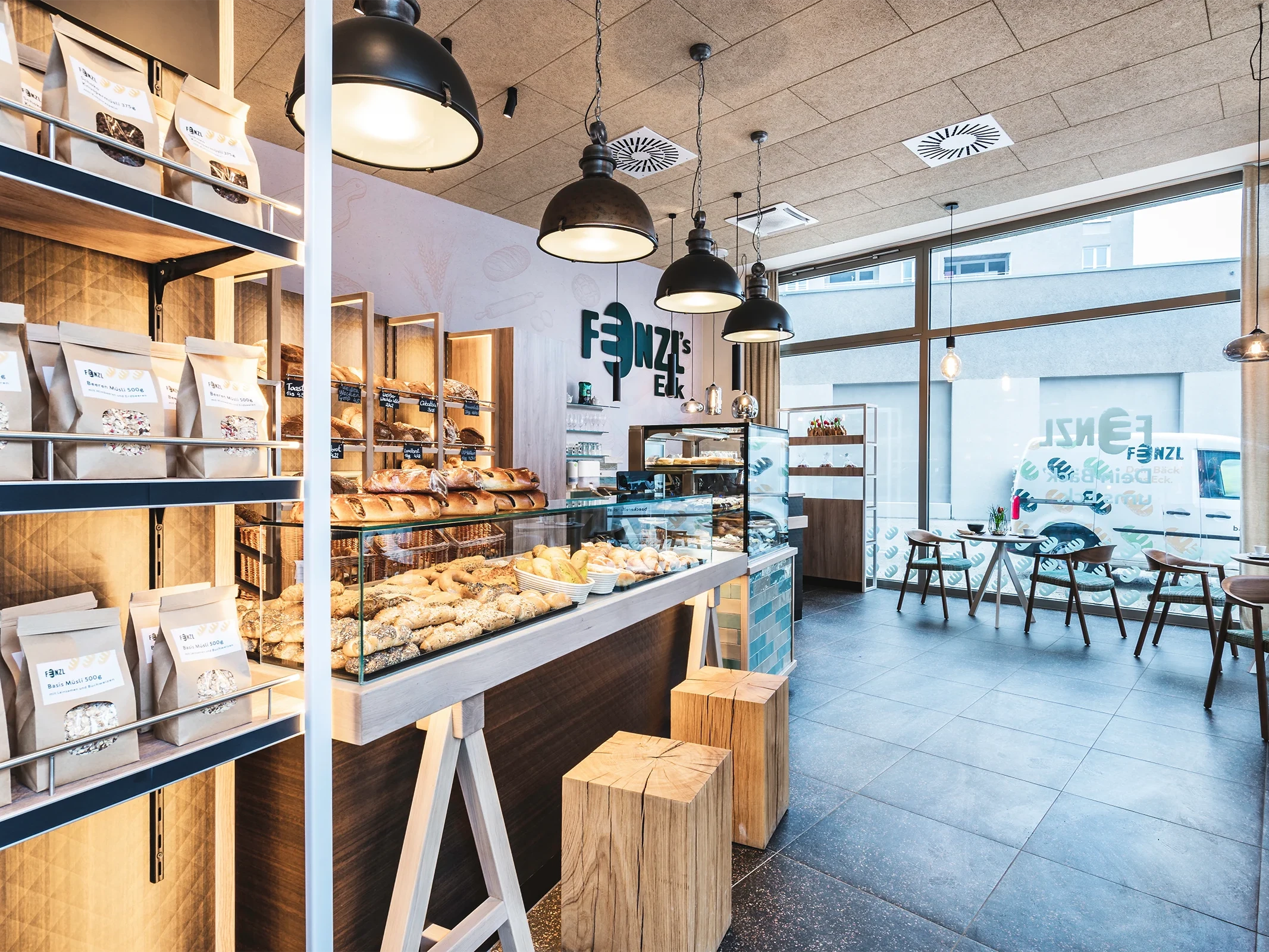 Gesamtansicht der Bäckerei mit beleuchtetem Brotregal, Glasvitrine mit Gebäck, naturbelassenen Holzklötzen als Stehhilfen und dem Café-Sitzbereich im Hintergrund.
