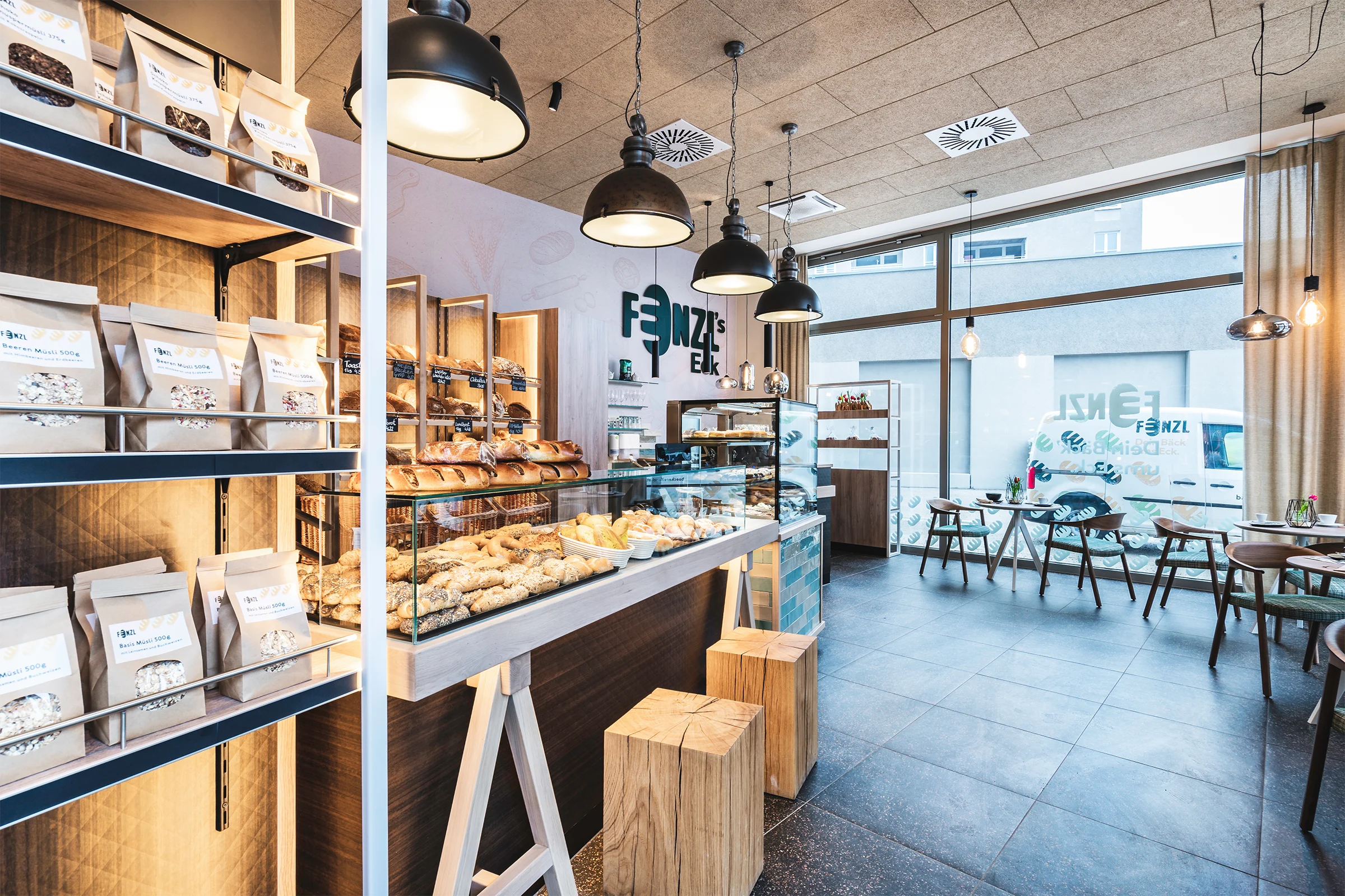 Gesamtansicht der Bäckerei mit beleuchtetem Brotregal, Glasvitrine mit Gebäck, naturbelassenen Holzklötzen als Stehhilfen und dem Café-Sitzbereich im Hintergrund.