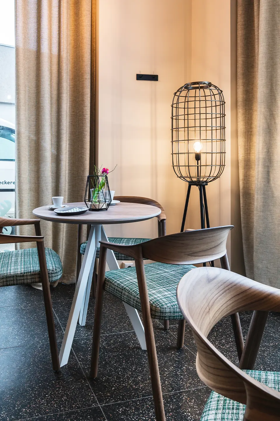 Gemütliche Café-Sitzecke mit Holzstühlen, grün-karierten Polstern, rundem Tisch und einer markanten Käfigleuchte auf Dreibeinstativ vor naturbeigen Vorhängen.