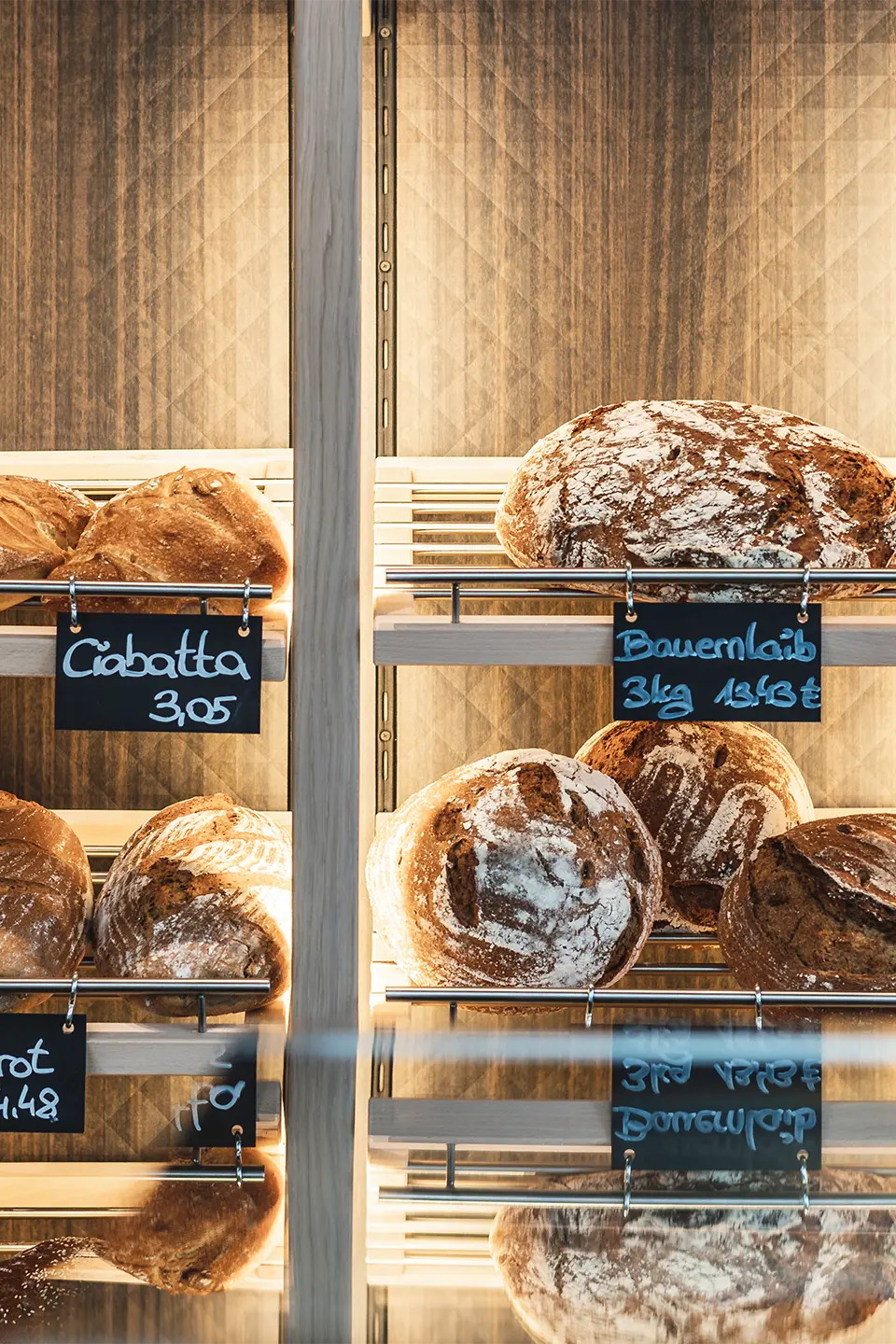 Nahaufnahme des hinterleuchteten Brotregals mit frischen Laiben – darunter Ciabatta und Bauernbrot – mit handgeschriebenen Kreidetafelschildern