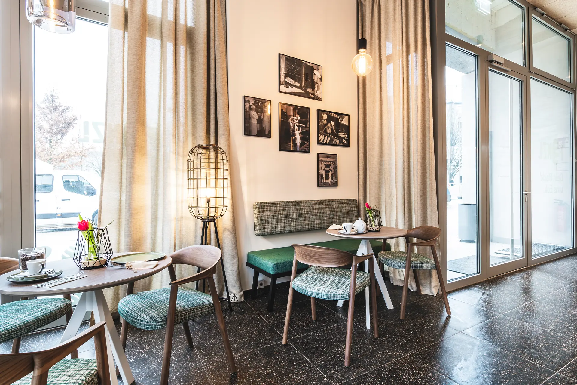 Heller Café-Sitzbereich mit grün-karierten Bänken und Holzstühlen, einer Käfigleuchte sowie einer Schwarz-Weiß-Fotogalerie aus der Bäckereigeschichte an der Wand
