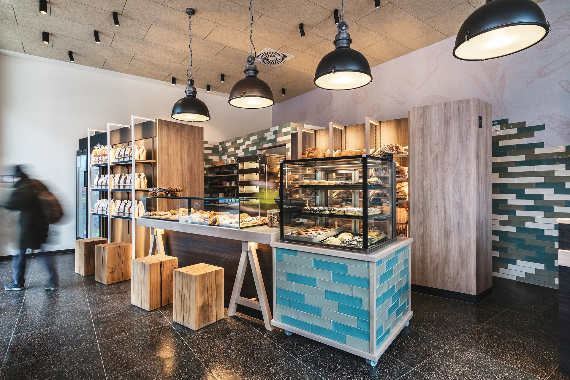 Verkaufstheke mit blauen Keramikfliesen, Gebäckvitrine und schwarzen Industriehängelampen vor einer gemusterten Fliesenwand in Grün- und Beigetönen