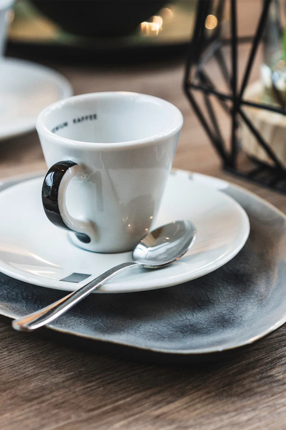 Detailaufnahme einer weißen Espressotasse mit schwarzem Henkel auf Untertasse und grauem Keramikteller, bereit für den Gast