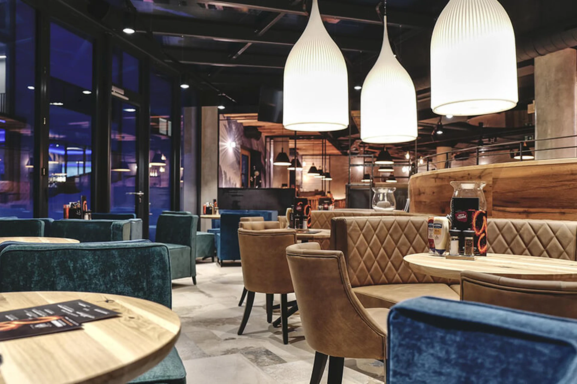 Stimmungsvoller Gastraum des Restaurants Gaspis Gerlos mit blauen und beigen Sesseln, runden Holztischen, großen weißen Hängelampen und Panoramafenster zur Skipiste.