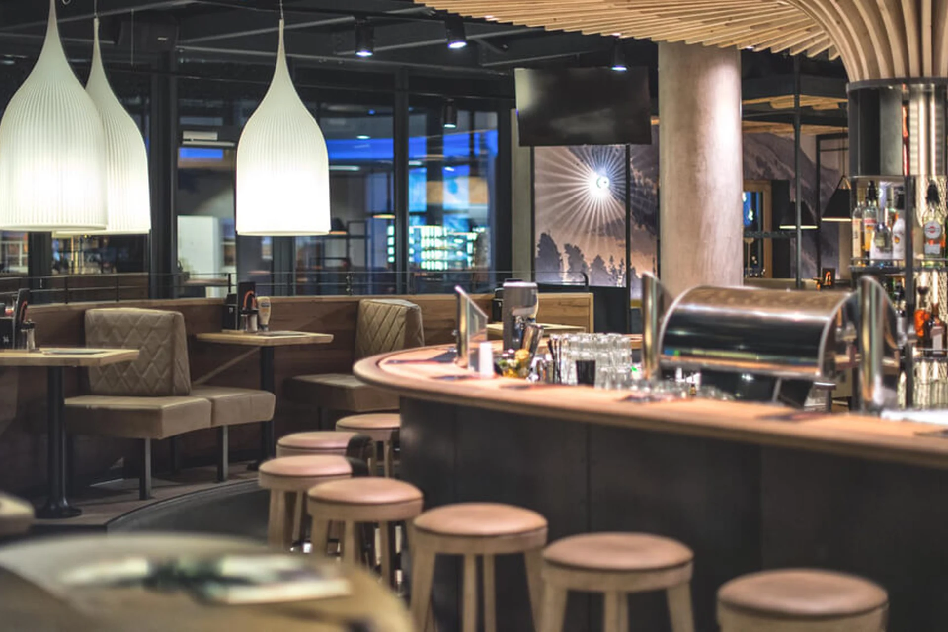 Rundtheke mit Barhockern im Restaurant Gaspis Gerlos mit weißen Hängelampen, beigen Sitzbänken und Bergpanorama-Wandbild im Hintergrund.