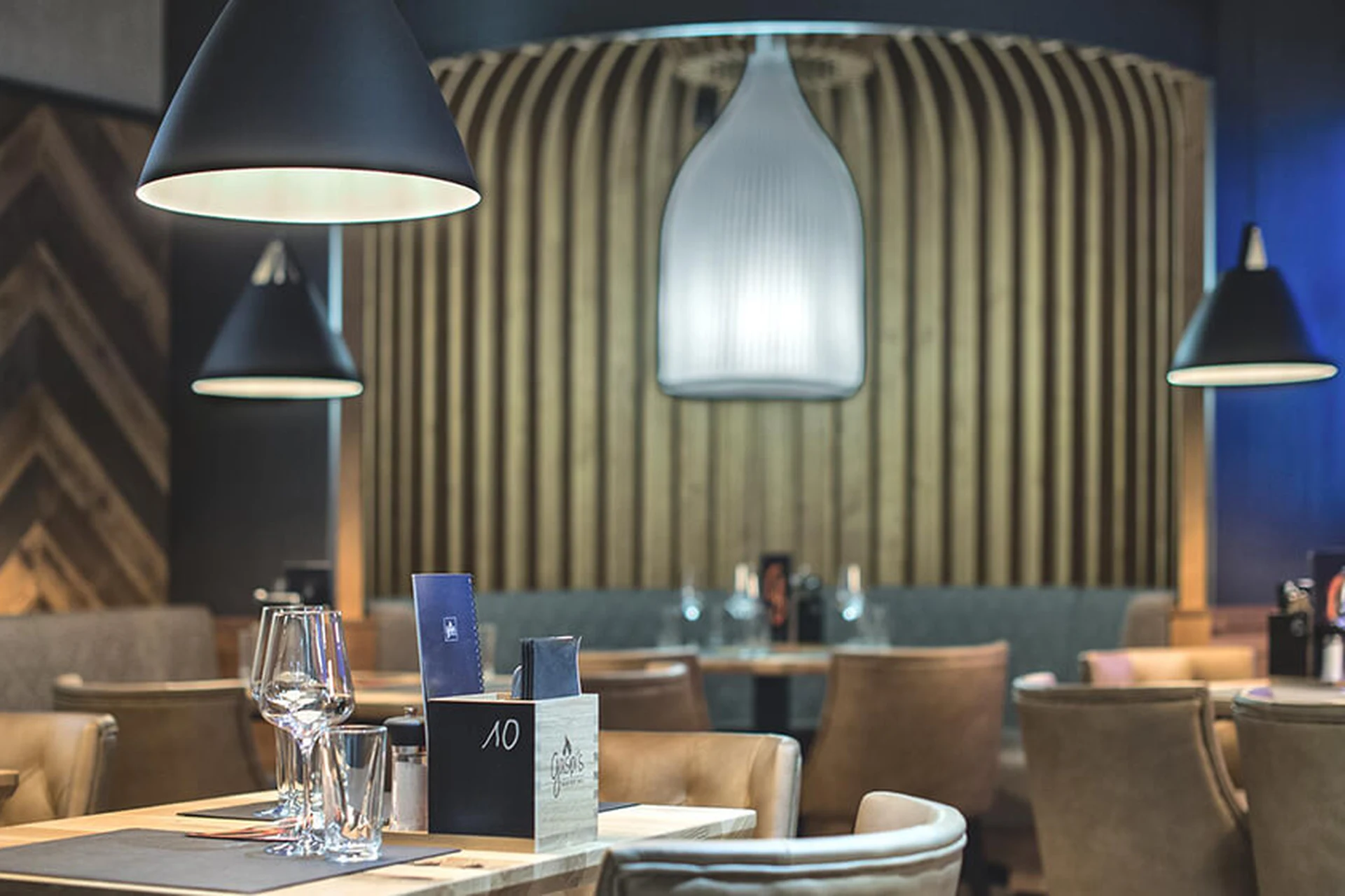 Gedeckter Tisch mit Weingläsern im Restaurant Gaspis Gerlos mit schwarzen und weißen Hängelampen, beigen Sesseln und gestalteter Wandvertäfelung.