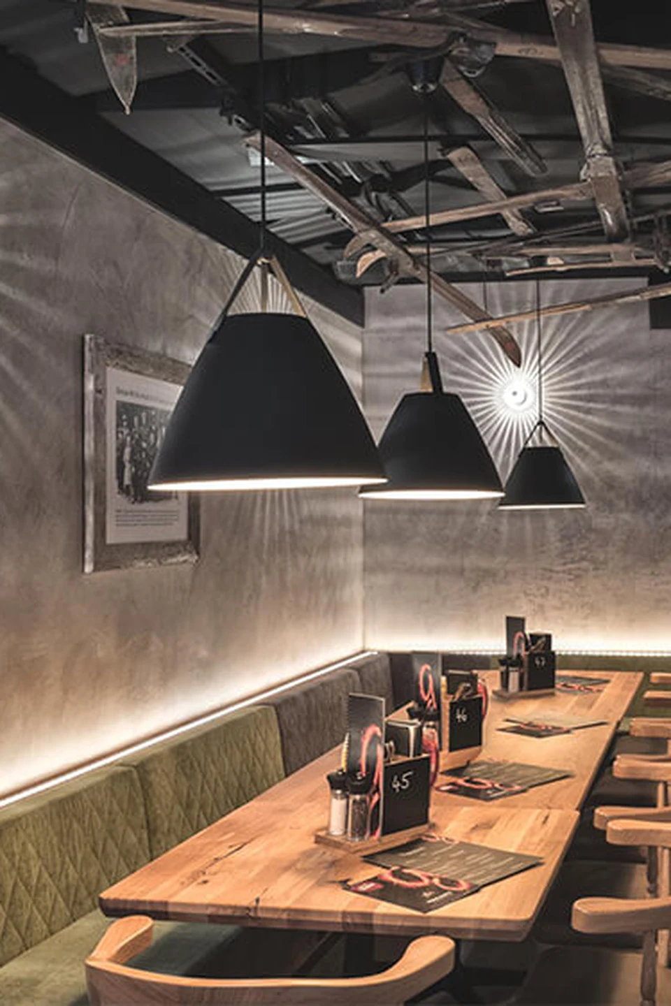 Separée im Restaurant Gaspis Gerlos mit langem Holztisch, grüner Sitzbank, schwarzen Hängelampen und rustikaler Betonwand mit indirekter Beleuchtung und an der Decke montieren vintage Ski.