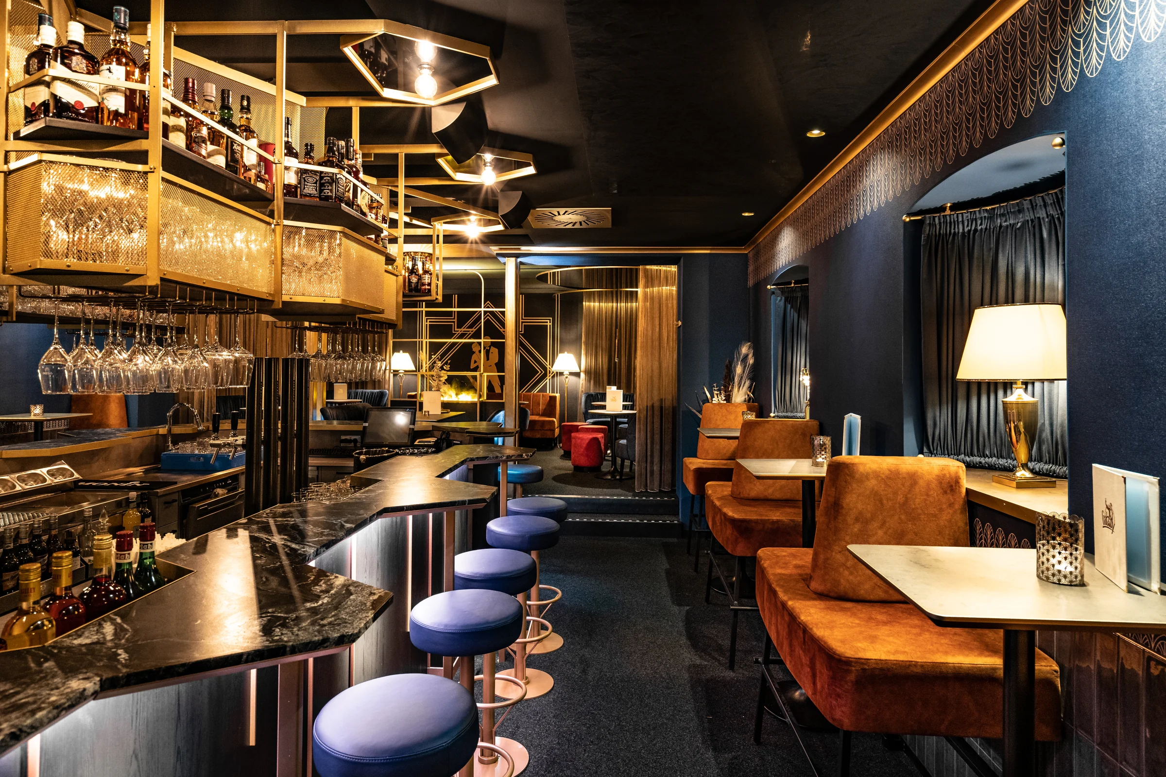 Elegante Art-déco-Bar mit goldenen Regalen, dunklen Wänden, blau gepolsterten Barhockern und schwarzem Marmortresen in stimmungsvoller Beleuchtung.